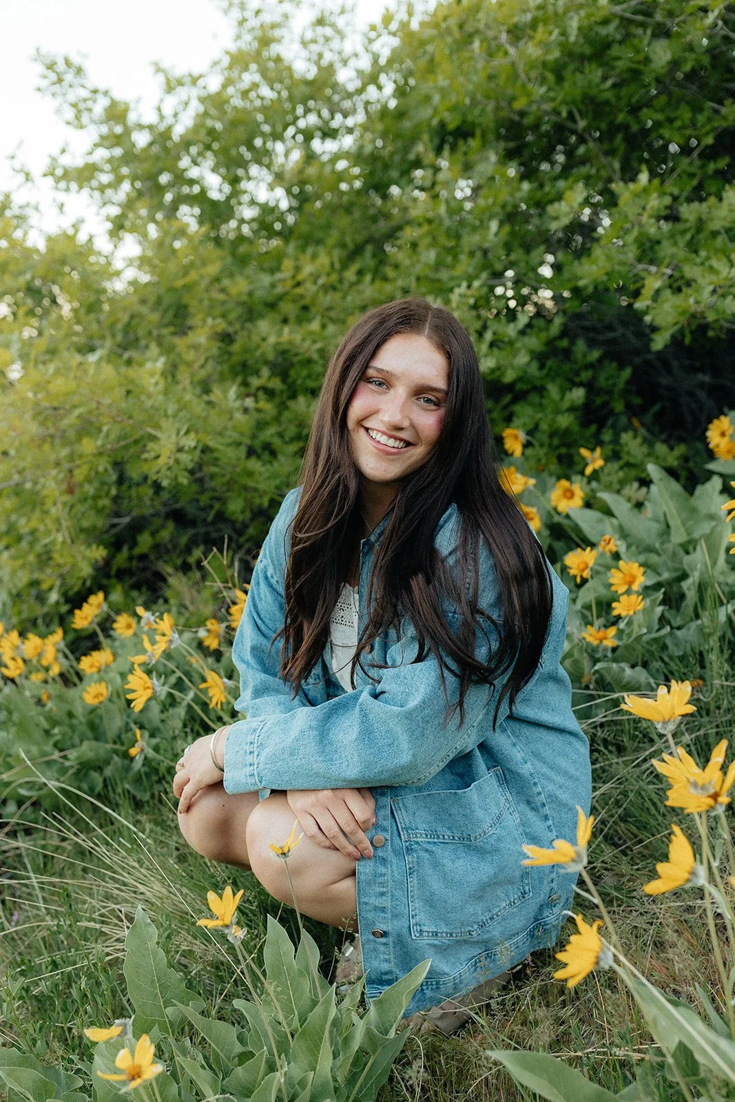 A young woman with long dark hair smiling and squatting among yellow flowers and green foliage outdoors.