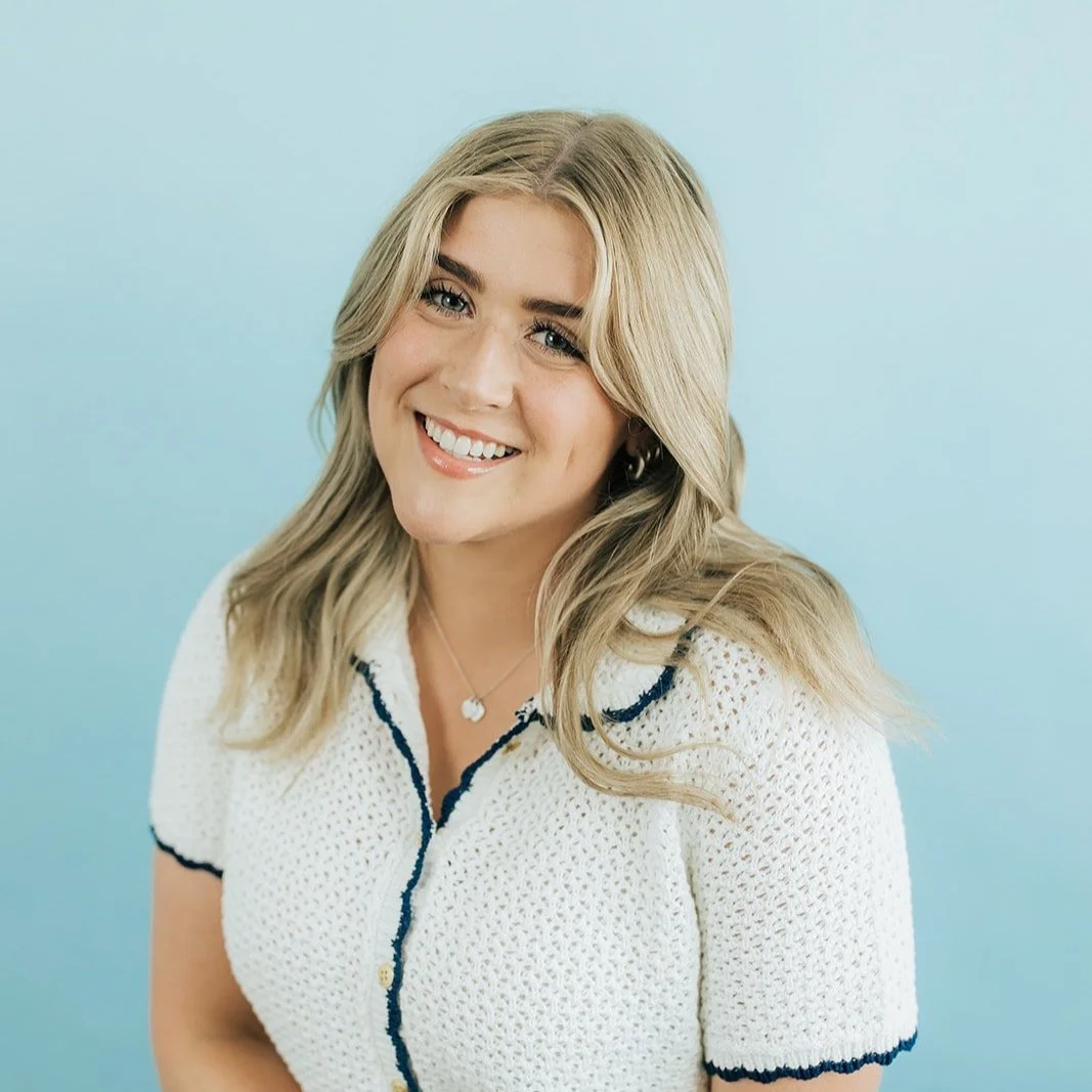 A smiling woman with blonde hair wearing a white short-sleeved knit top, standing against a light blue background.