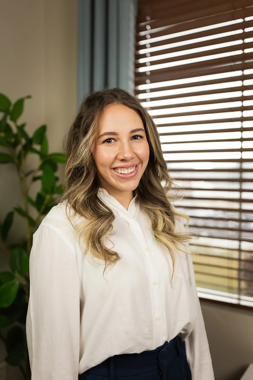 A smiling young woman with wavy brown hair wearing a white blouse standing in front of a window with brown blinds and a green houseplant.