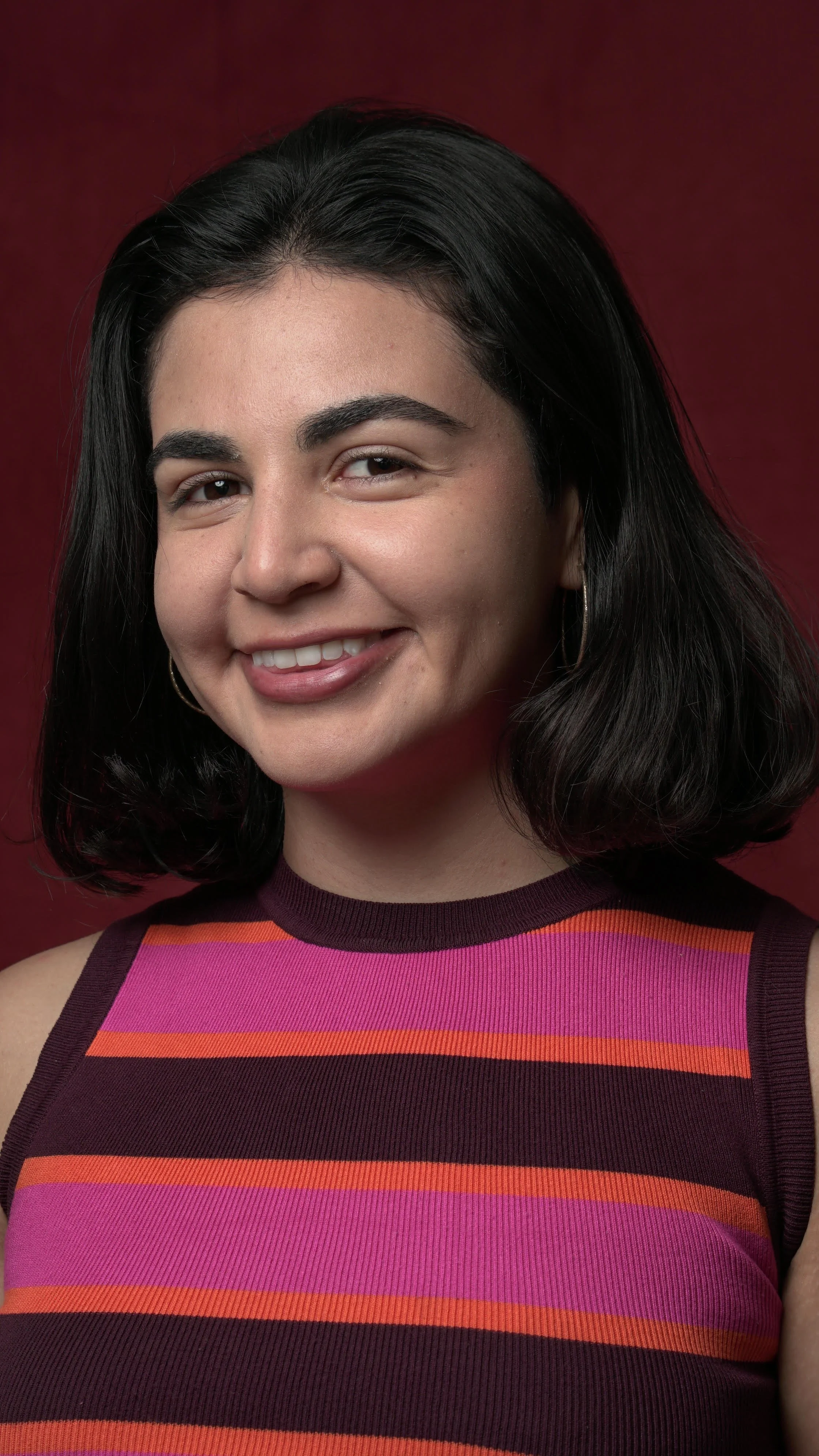 A headshot of a woman wearing a pink, orange, and brown striped top against a burgundy background.