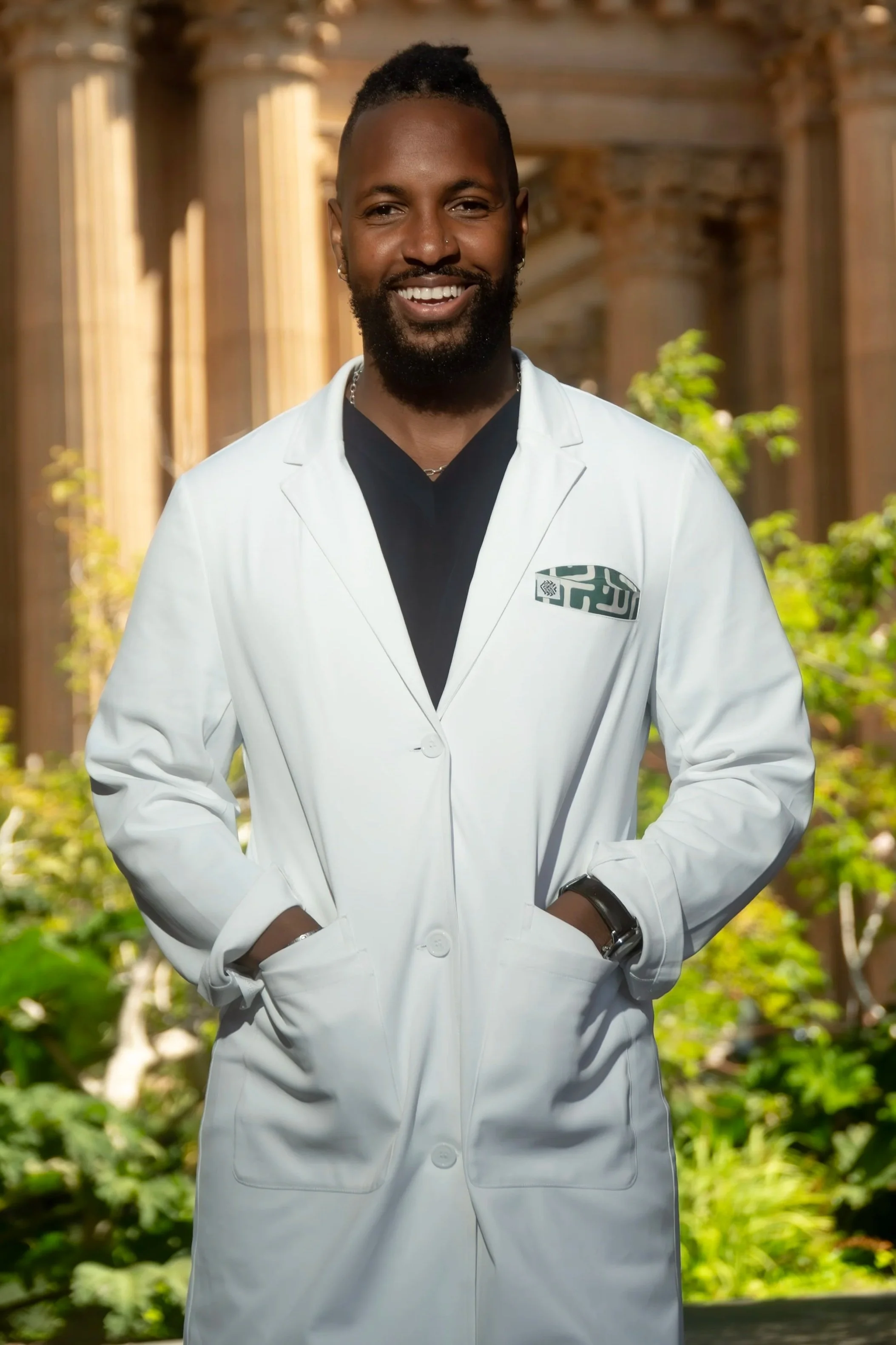 A headshot of a man wearing a white coat with black scrubs underneath. His hands are inside the pockets of the white coat against the Palace of Fine Arts.