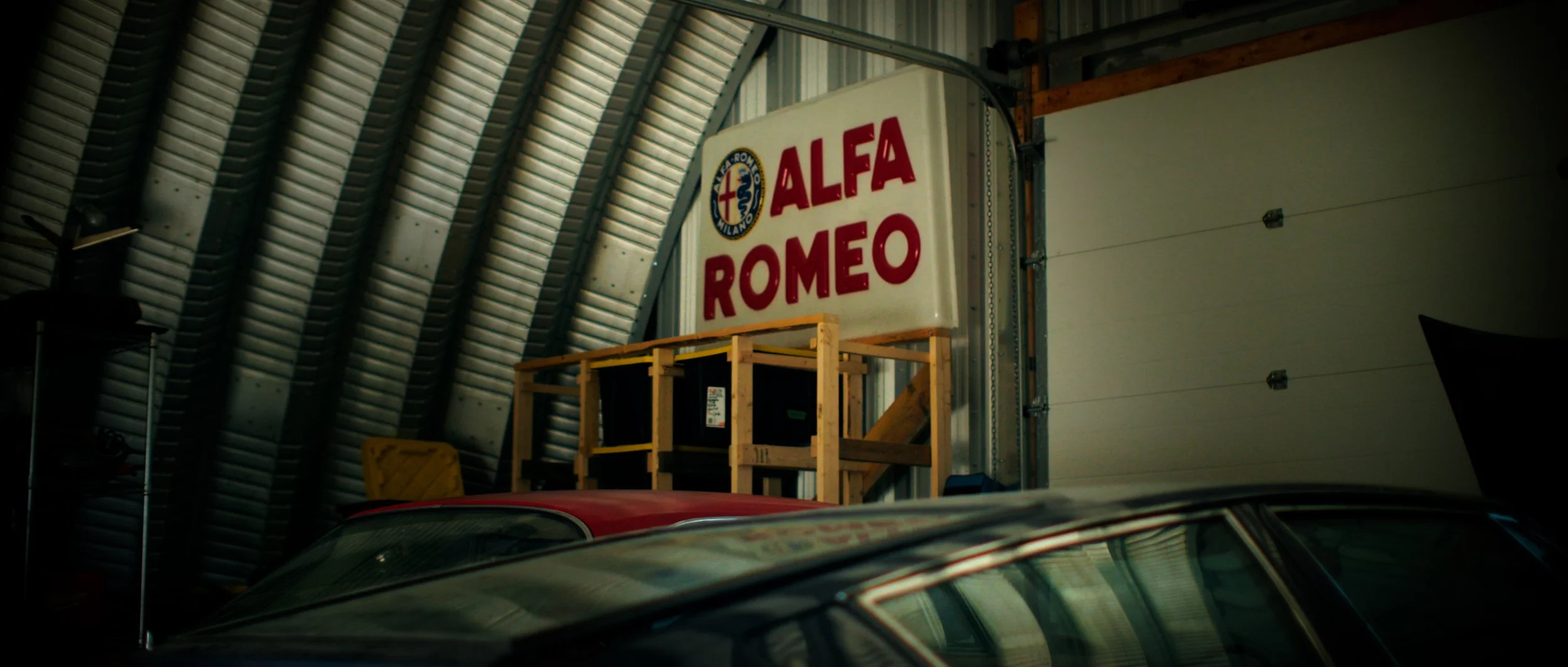 Indoor storage area with a large sign that reads 'Alfa Romeo' and features the Alfa Romeo emblem. The sign is mounted on a wall or structure with visible metal beams, and there is a wooden frame supporting some items below it. A portion of a car's ro