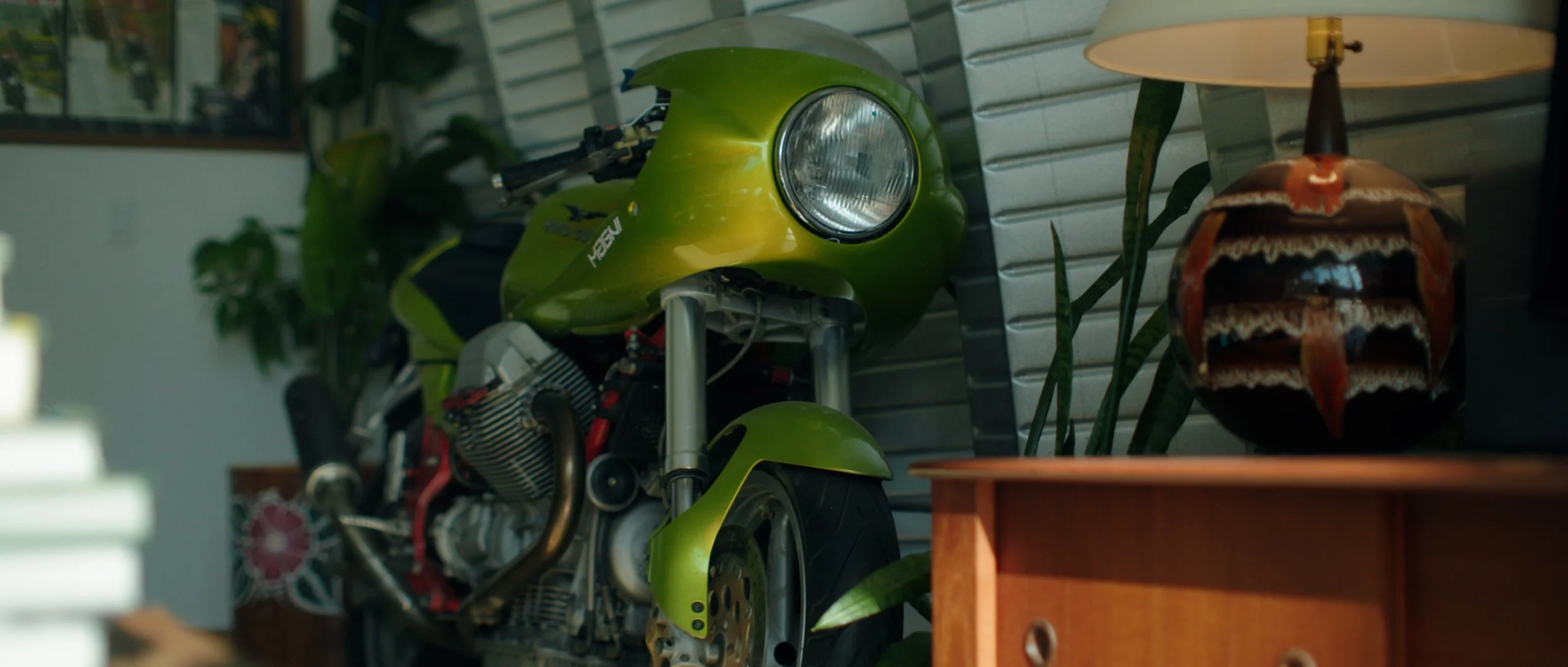 A green motorcycle with a round headlight parked indoors near a wall and a vintage lamp on a wooden table.