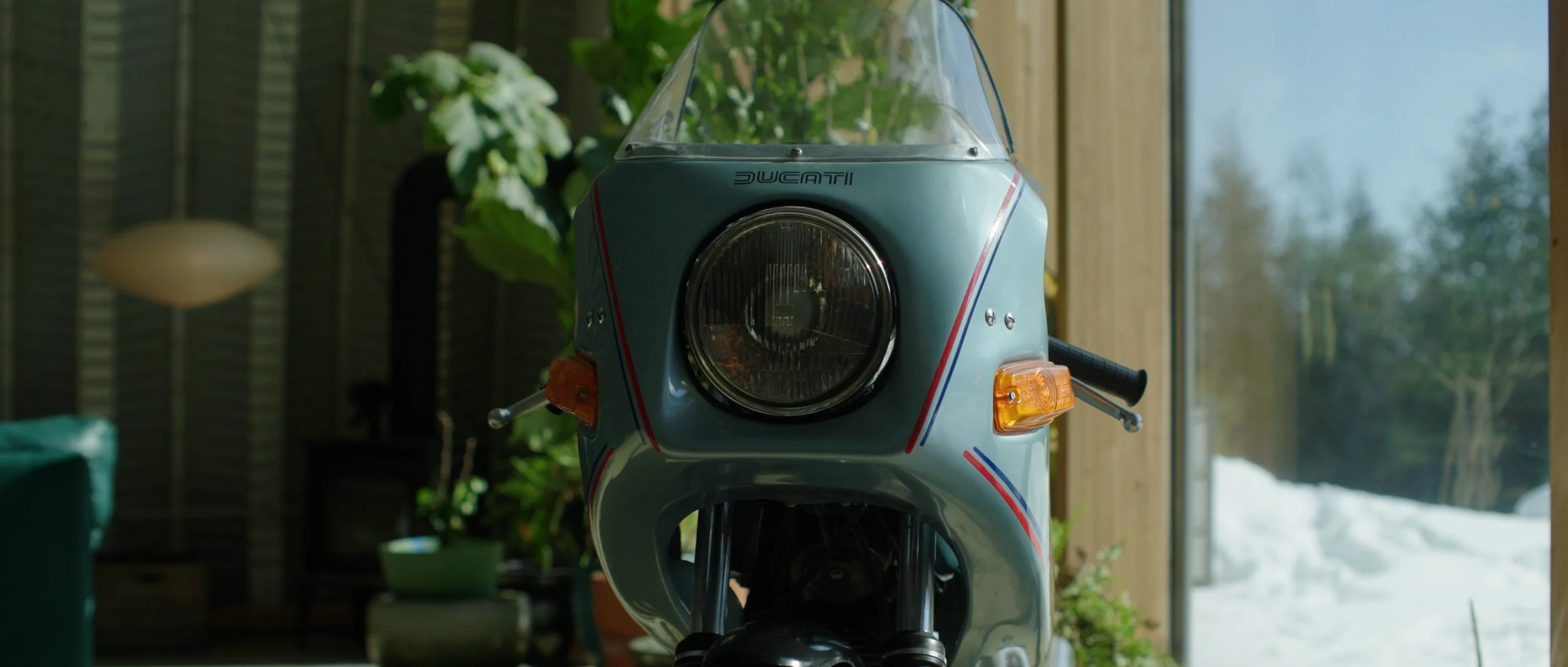Front view of a vintage light blue Ducati scooter with a windshield indoors near a window with a snowy landscape outside.