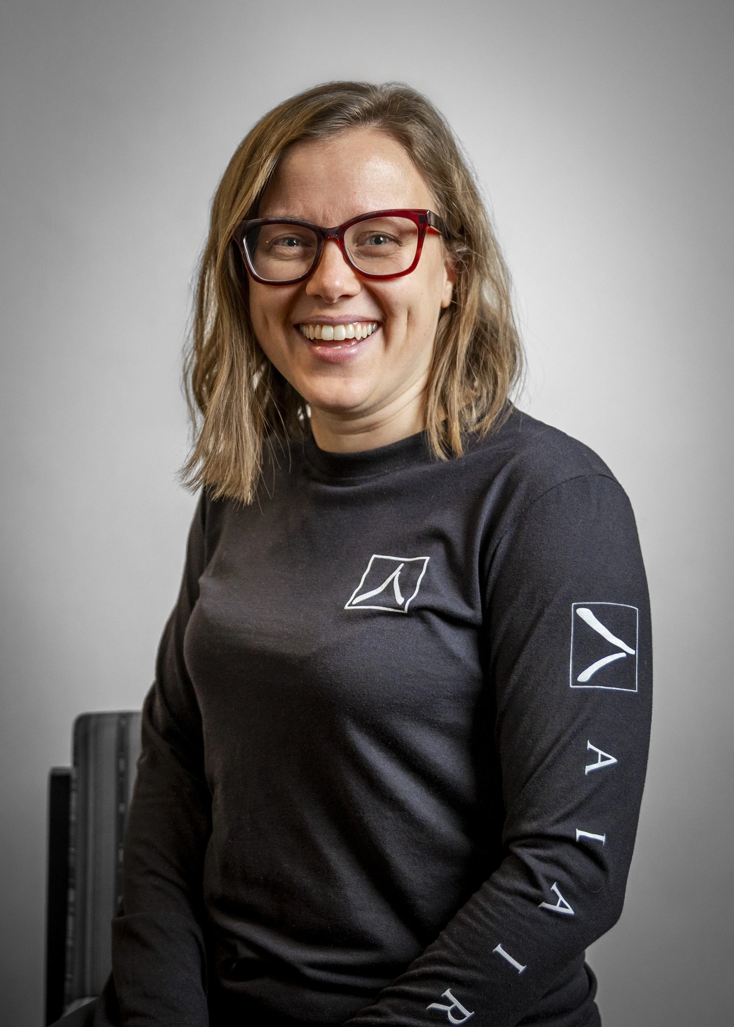 A woman wearing red glasses and a black long-sleeve shirt with white logo and text, smiling at the camera against a gray background.