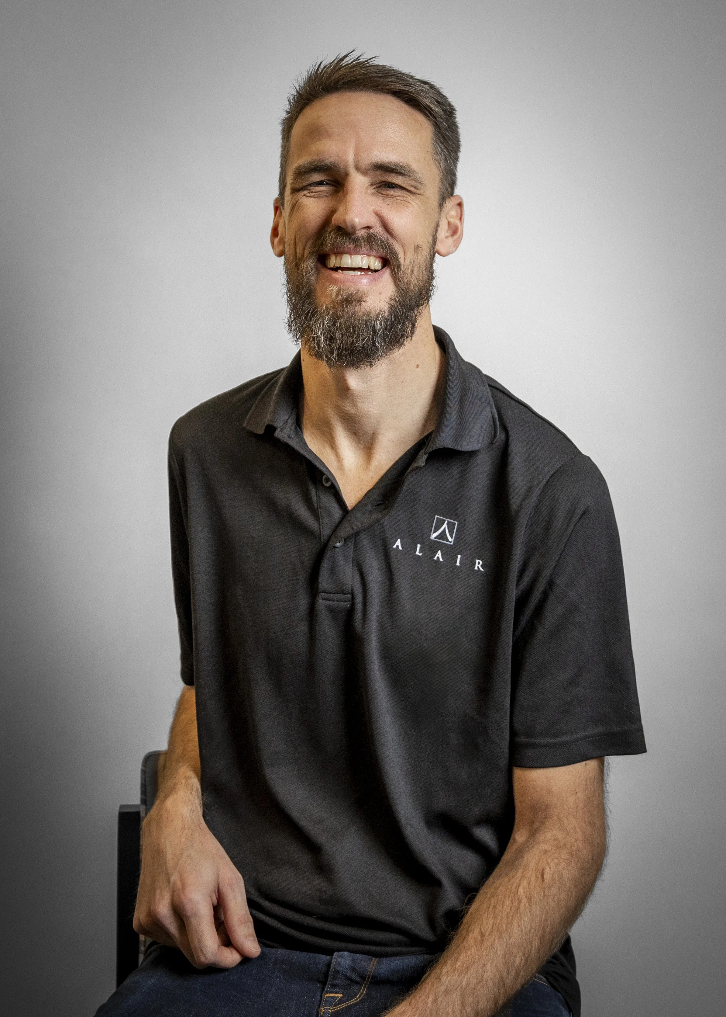 A man with a beard and mustache, wearing a black collared shirt with a logo and the word 'ALLA IR', sitting and smiling against a neutral background.