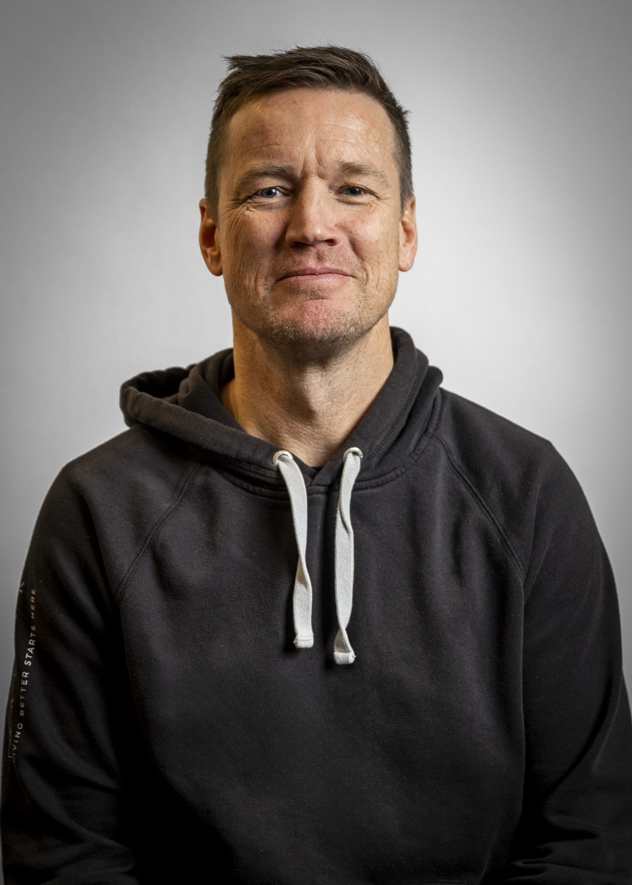 A middle-aged man with short brown hair and light skin, wearing a black hoodie with white drawstrings, looking at the camera with a slight smile against a plain gray background.