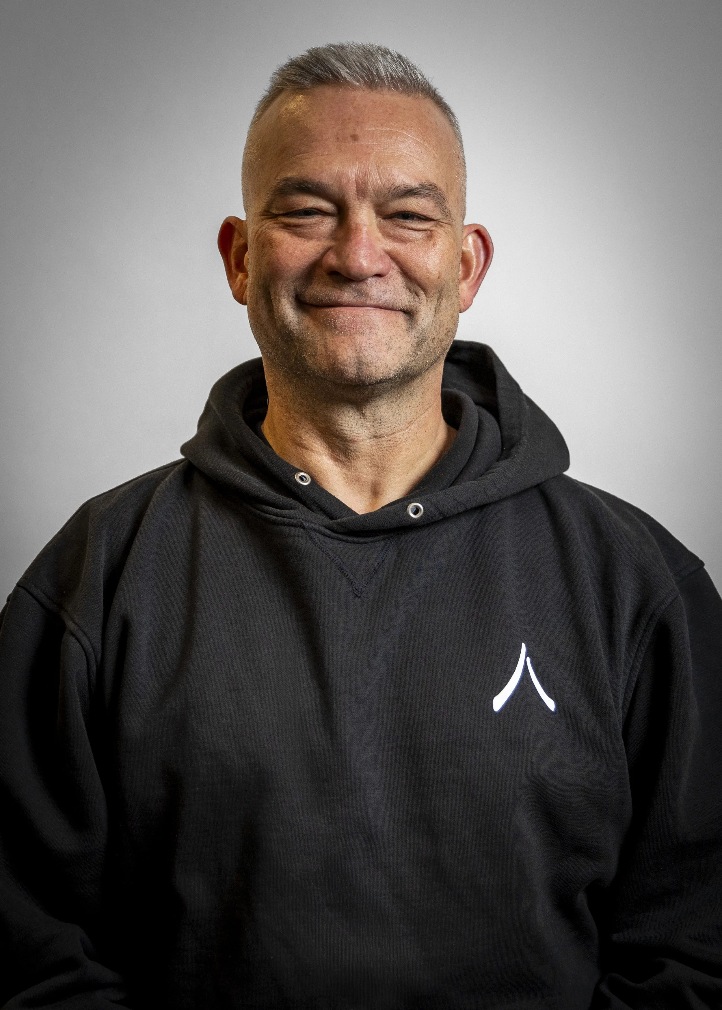 A smiling man with short gray hair wearing a black hoodie with a logo on the chest, standing against a plain gray background.