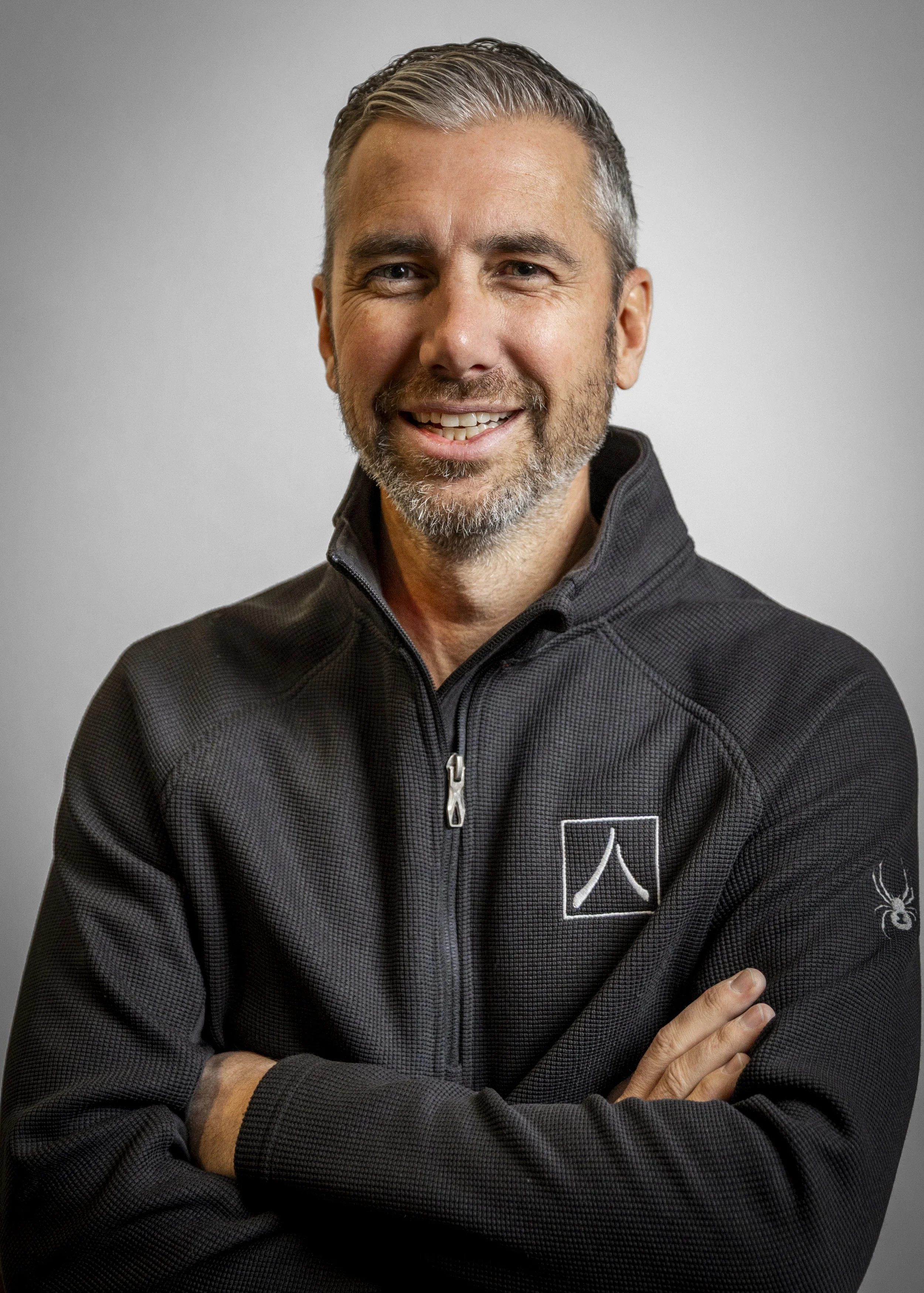 A smiling middle-aged man with gray hair and a beard, wearing a black zip-up jacket with a white logo and a spider logo on the sleeve, standing with arms crossed against a plain gray background.