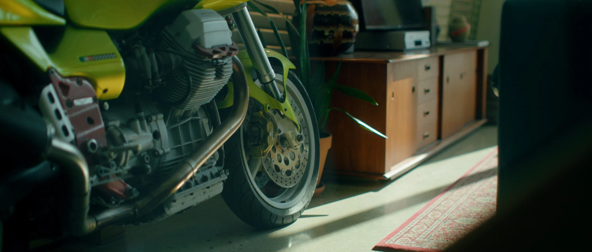 A motorcycle in a room with a wooden cabinet, potted plants, and a patterned rug on the floor.