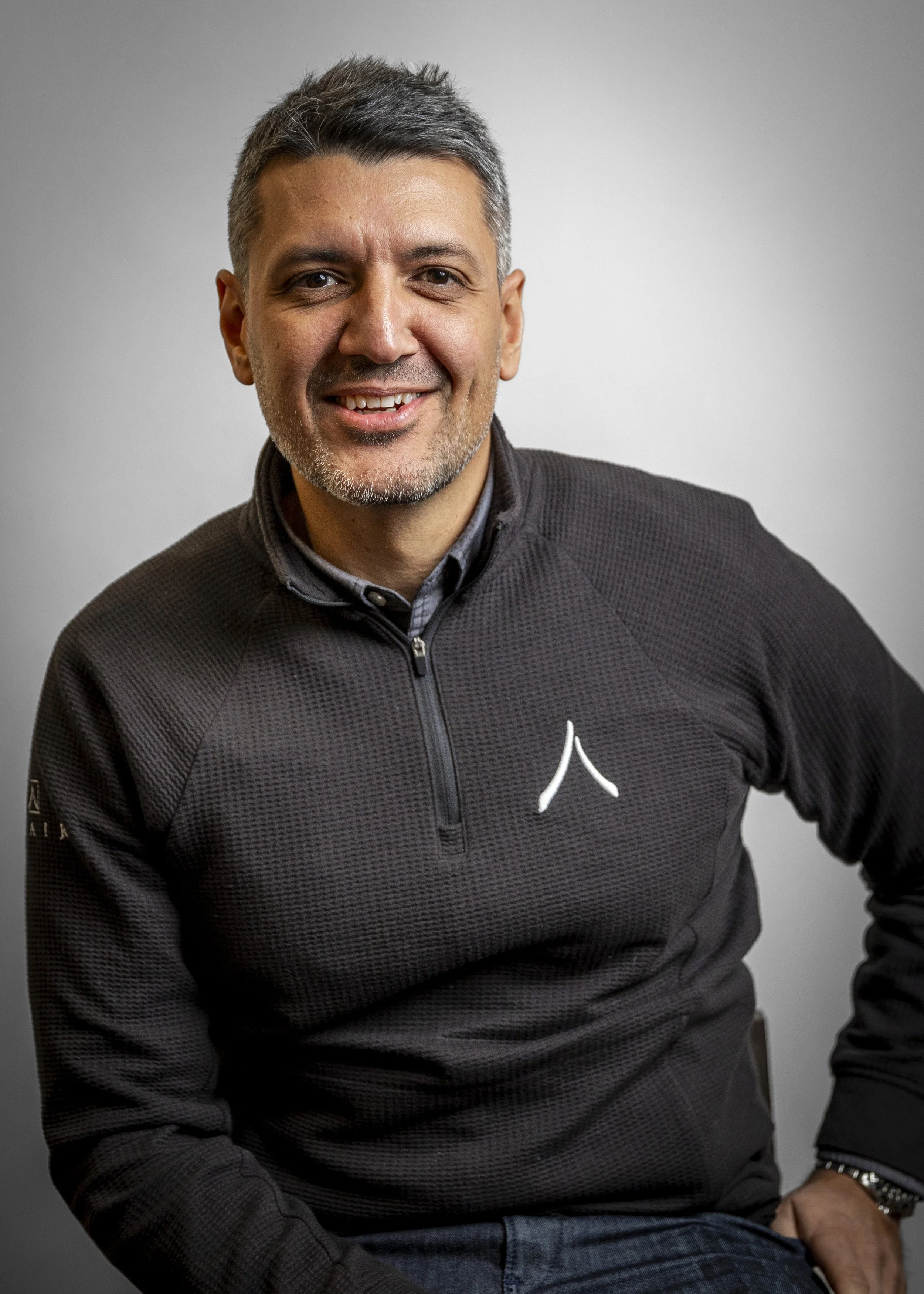 A smiling middle-aged man with short gray hair and stubble, wearing a black zip-up athletic jacket with a white logo on the left chest, sitting against a plain gray background.