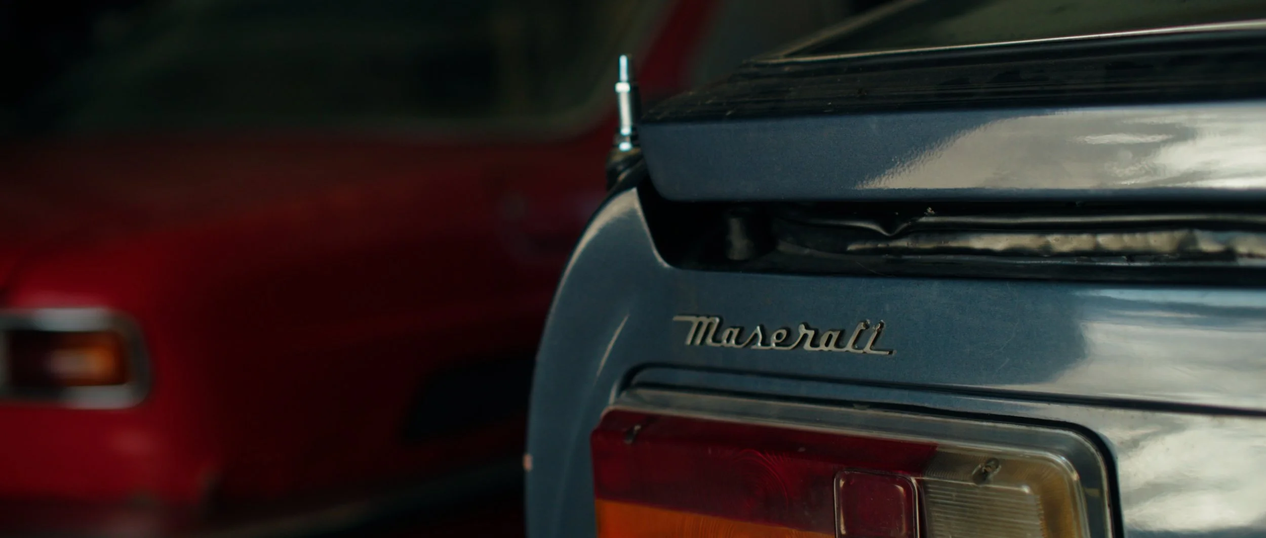 Close-up of the rear end of a silver Maserati car, focusing on the Maserati emblem with other vehicles blurred in the background.