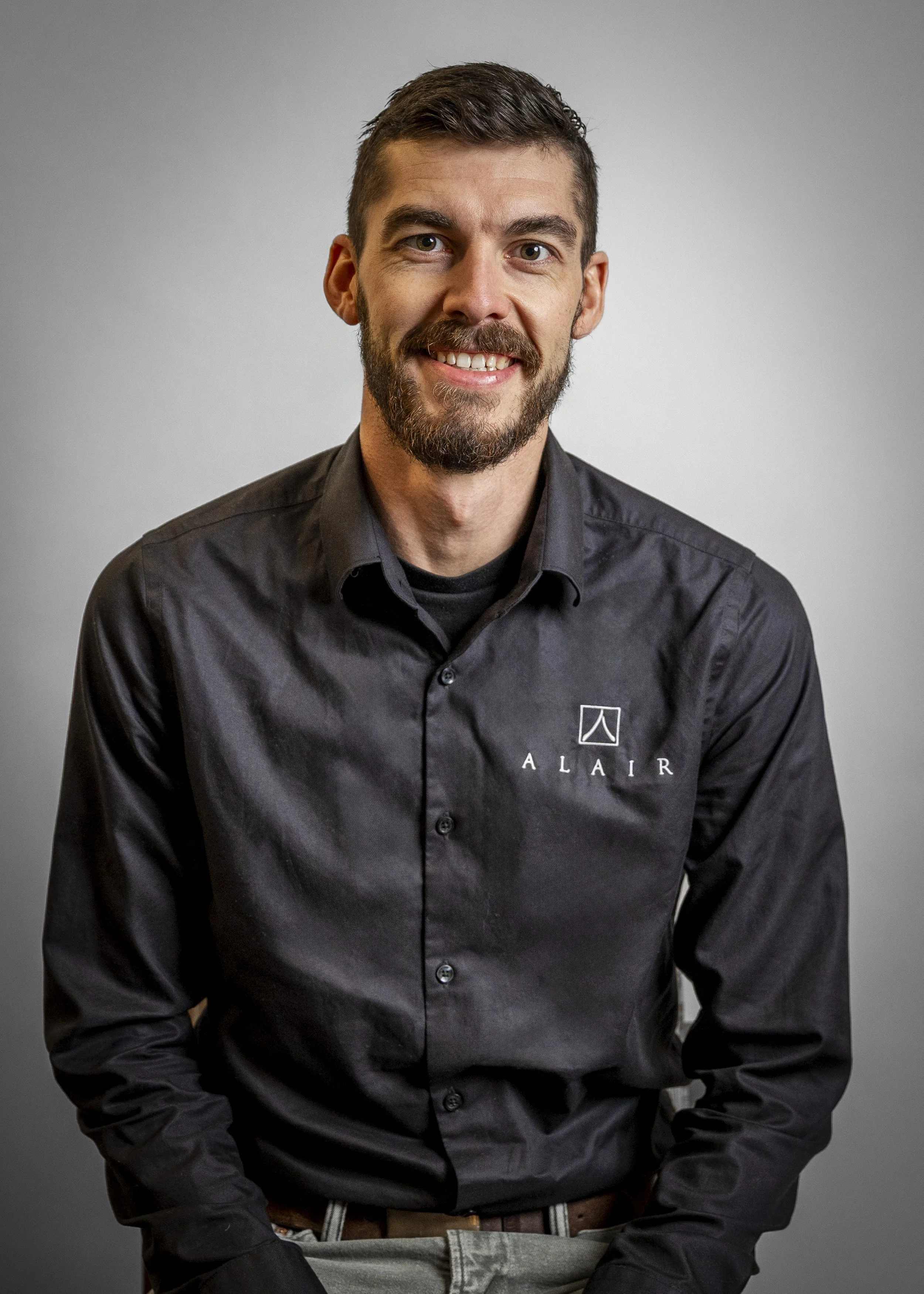 A man with dark hair and a beard wearing a black button-up shirt with a logo and the word 'ALAIR' on it, smiling at the camera against a gray background.