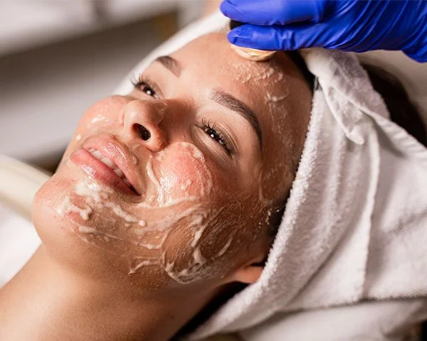 Una donna riceve un trattamento di pulizia del viso, con crema sul volto e un'operatore che indossa dei guanti blu che applica il prodotto.