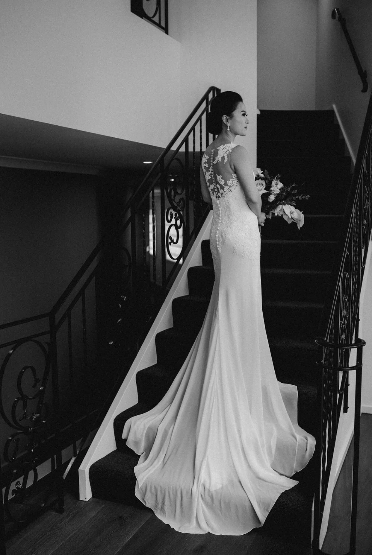 Black and white photo of a bride in a wedding dress with a train, holding a bouquet, standing on a staircase.