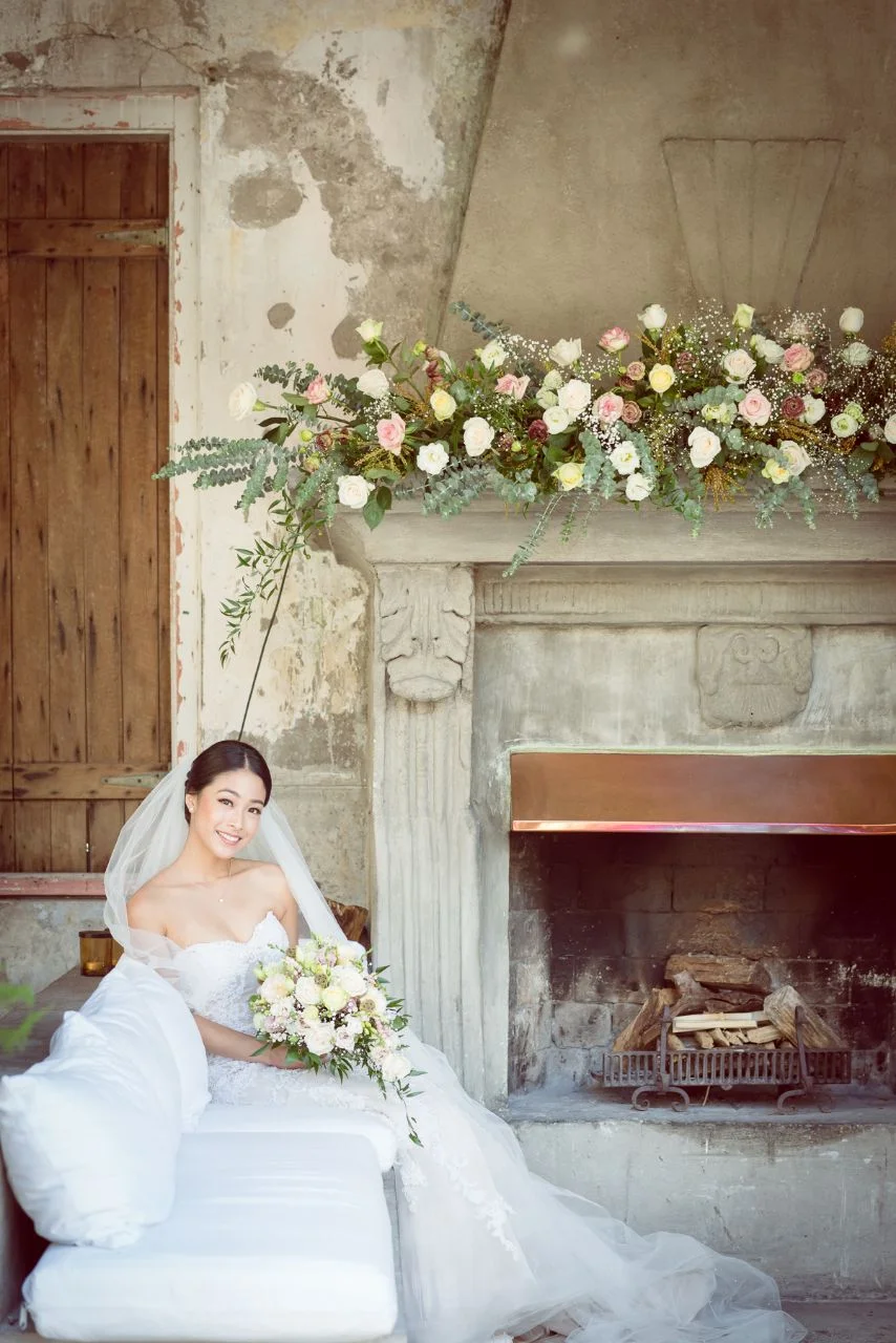 A bride in a white wedding dress and veil sitting on a white couch, holding a bouquet of flowers, next to a stone fireplace decorated with a large floral arrangement.