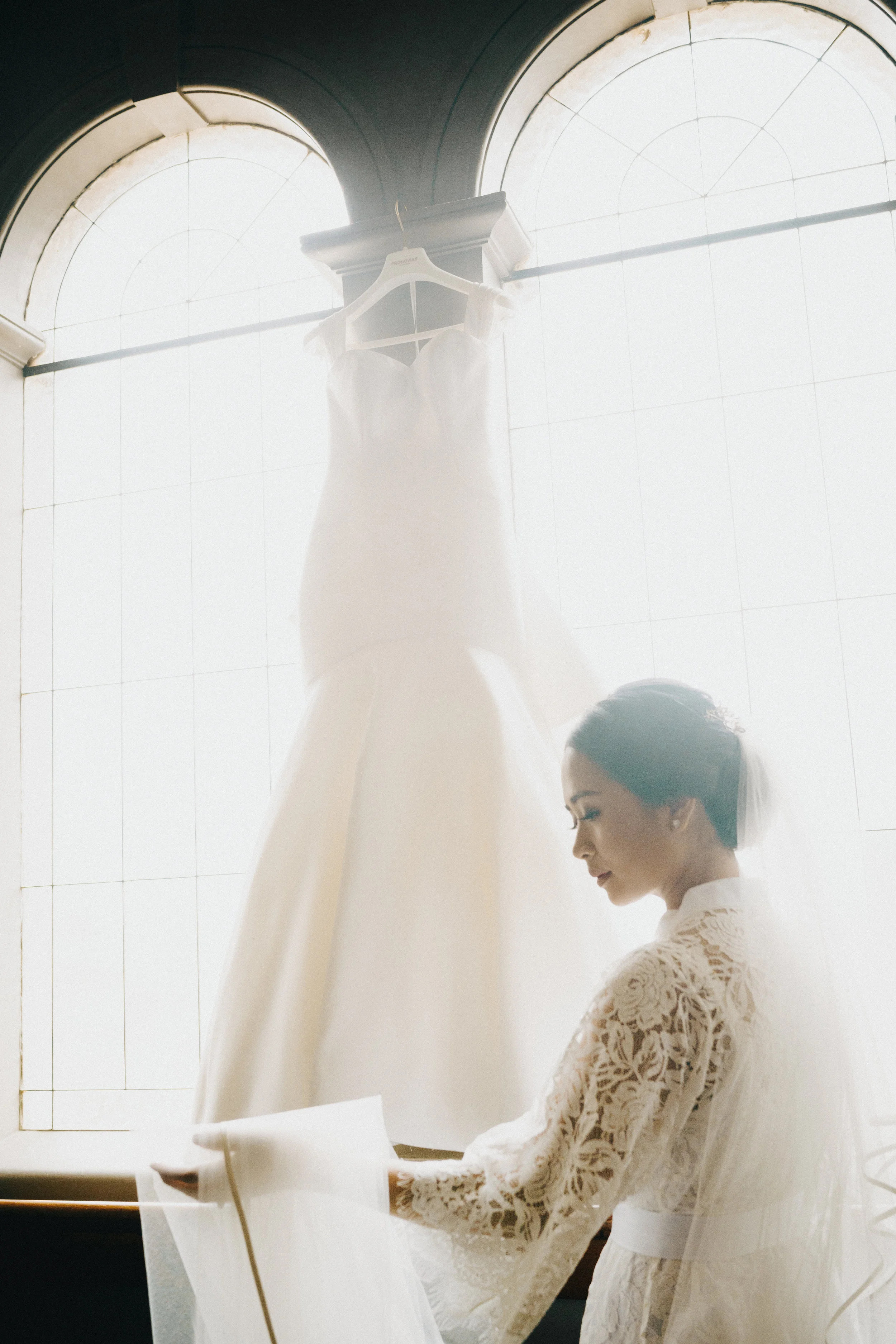 A woman in bridal attire reading under a hanging wedding dress in front of a large, bright window.