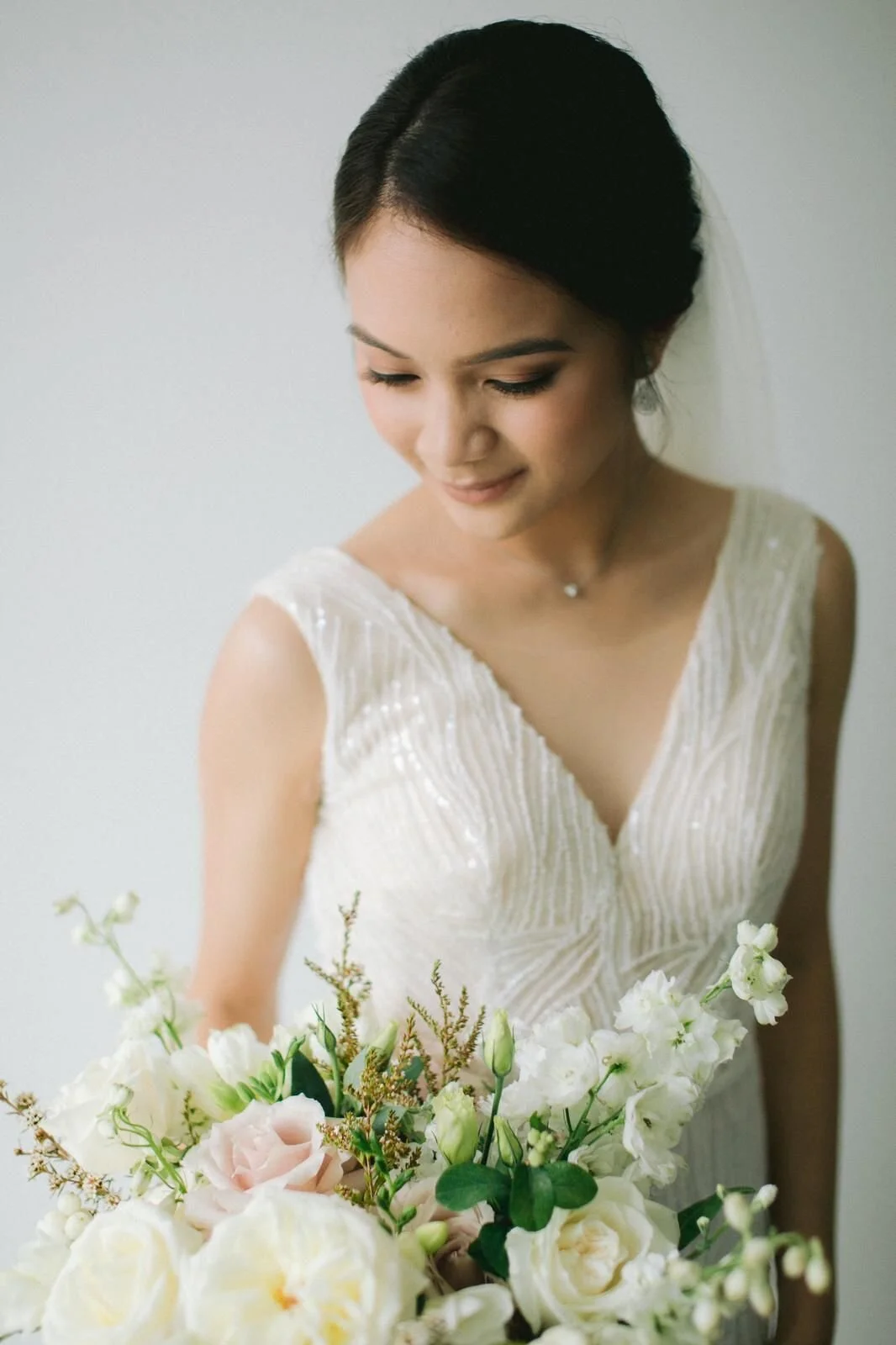 A bride in a white dress holding a bouquet of white and blush pink flowers, with her eyes downcast and a gentle smile.
