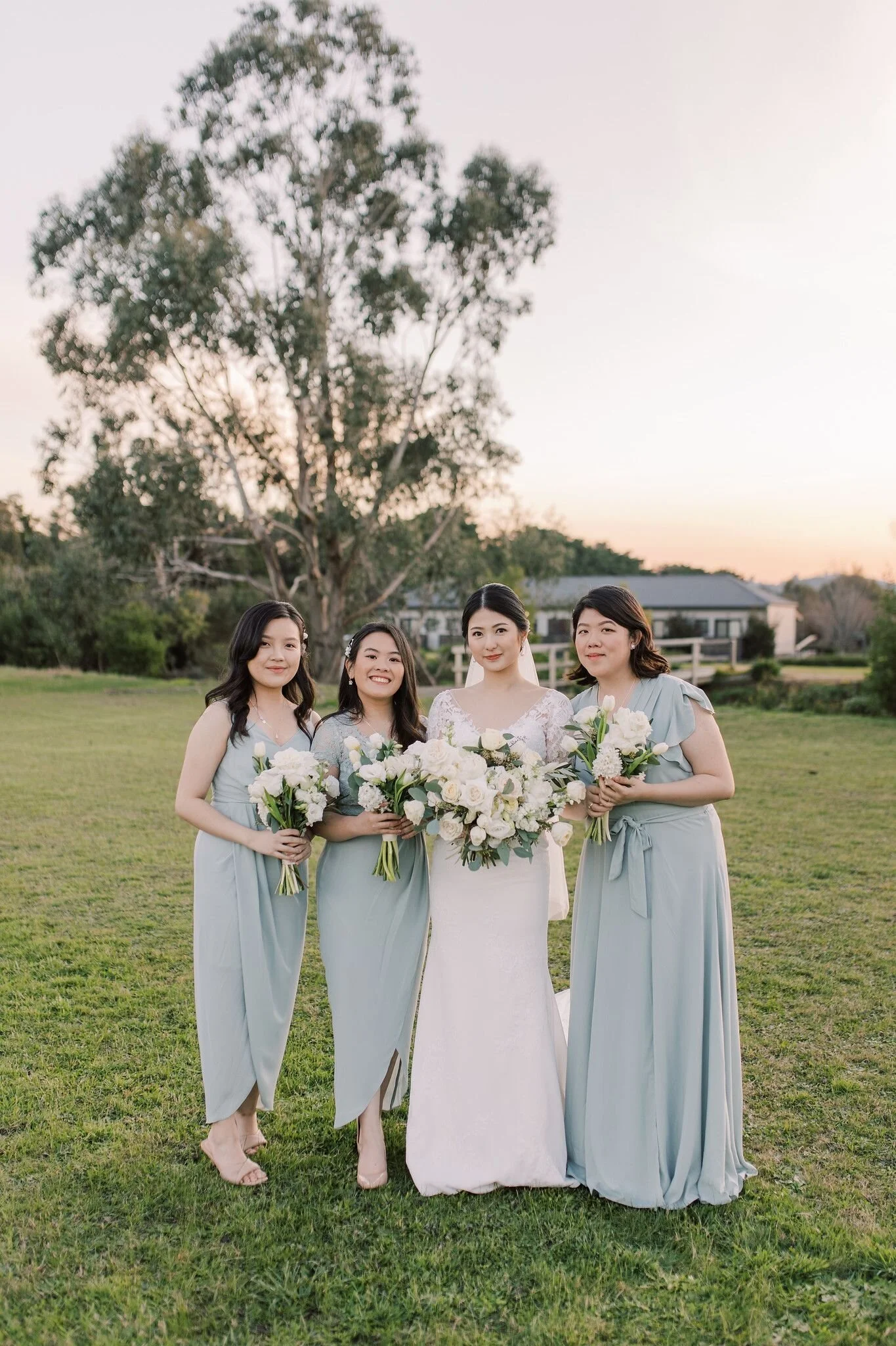 A bride and three bridesmaids standing outdoors on grass, holding bouquets of white flowers, during sunset or early evening.