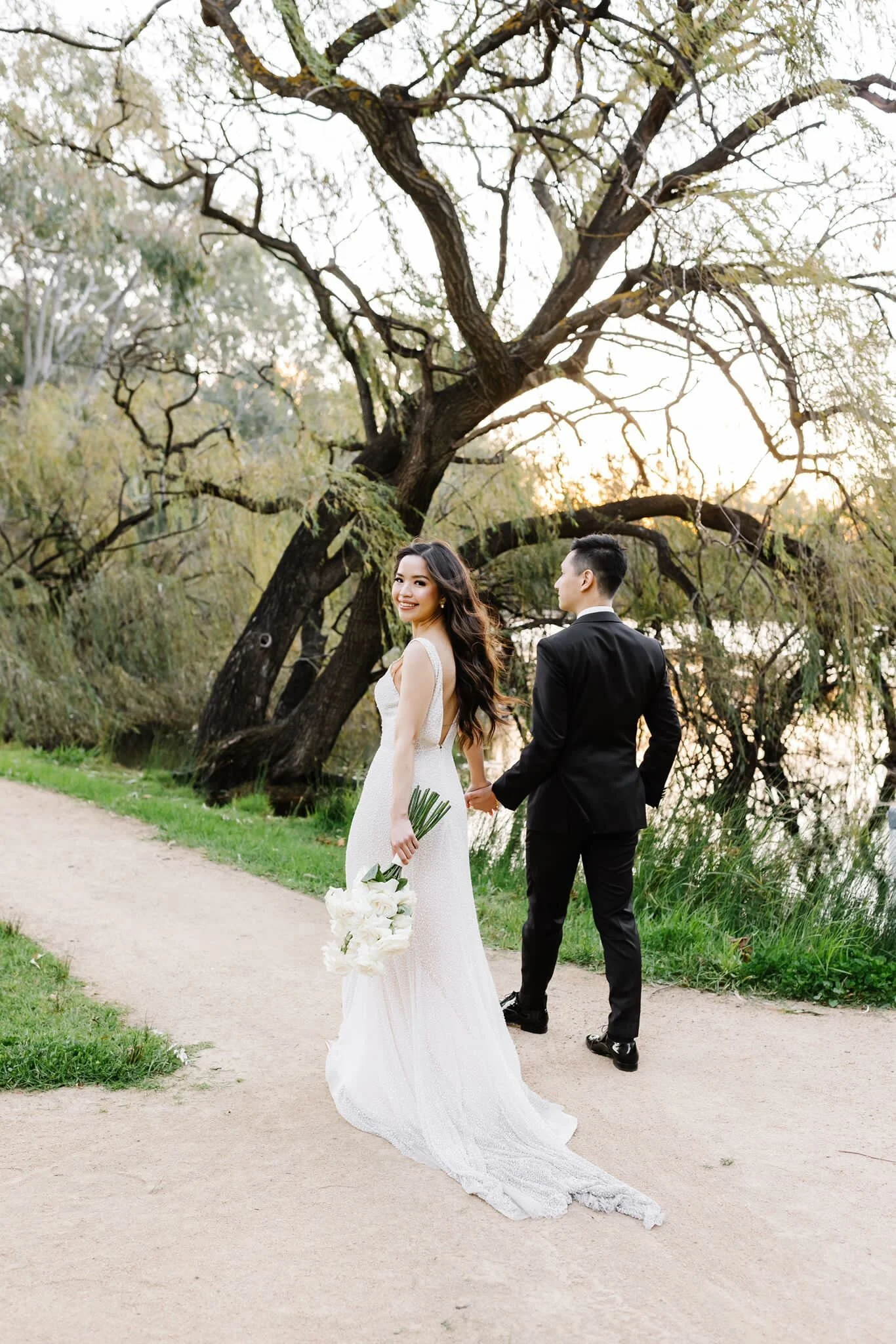 A bride and groom holding hands on a dirt path by a river during sunset. The bride is smiling and holding a bouquet of white flowers. The groom is facing away from the camera, wearing a black suit, while the bride is wearing a white wedding gown.