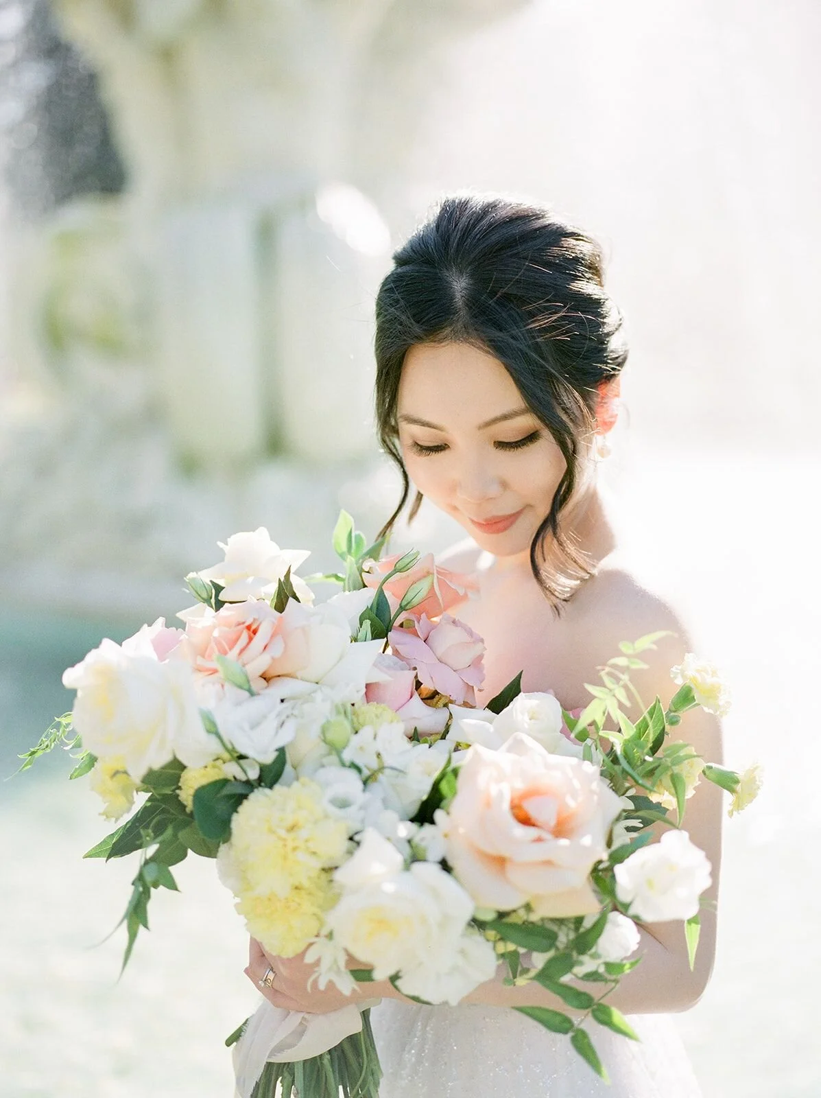 A woman in a wedding dress holding a large bouquet of white, pink, and yellow flowers, with her eyes closed and a gentle smile.
