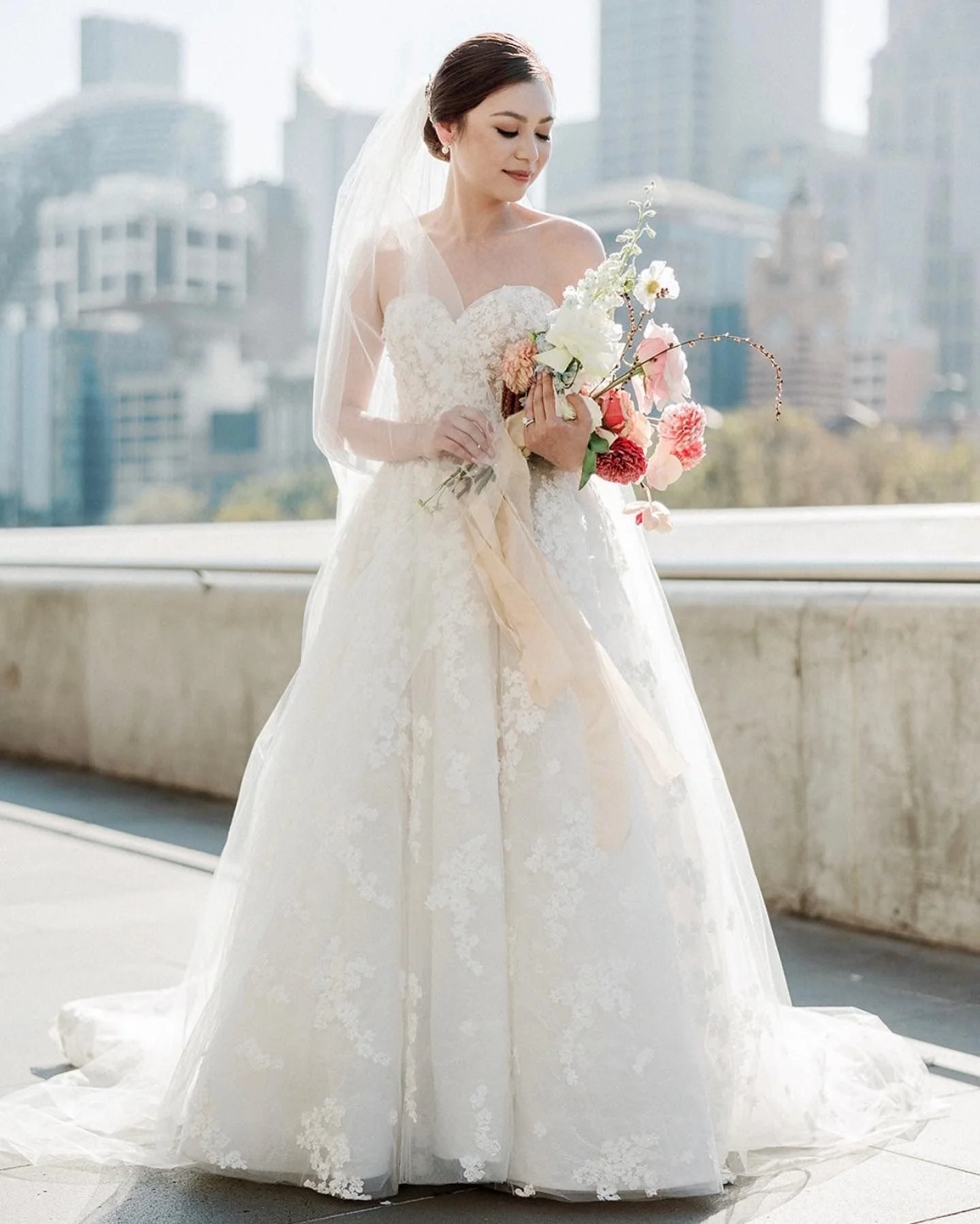 Bride in a lace wedding gown holding a bouquet of colorful flowers, standing outdoors with city buildings in the background.