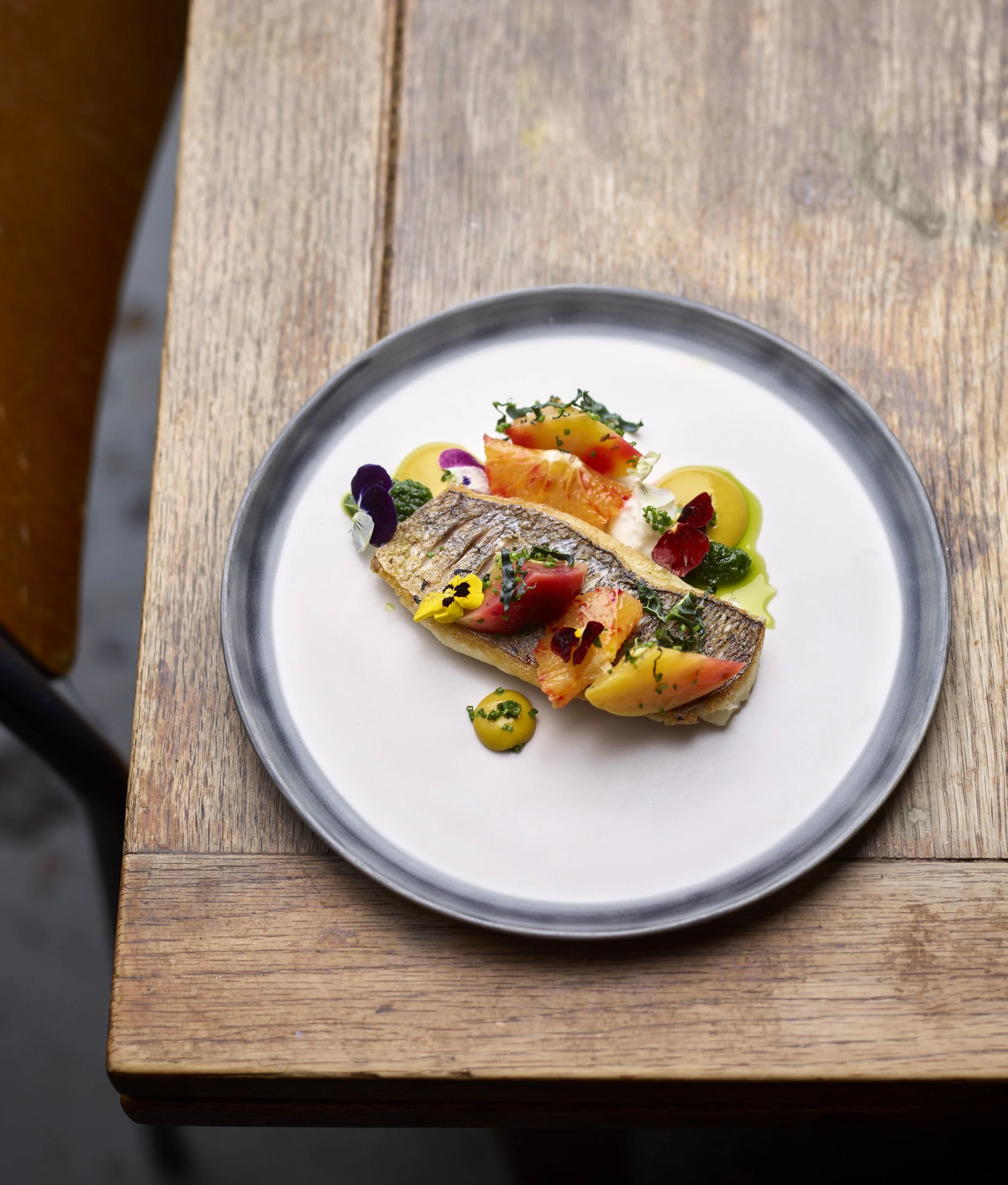 Close-up of a plated dish with fish, colorful vegetables, herbs, edible flowers, and drizzles of yellow sauce on a white plate, resting on a wooden surface.