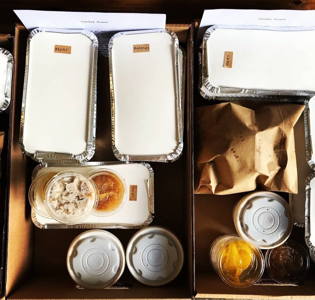 Takeout food containers labelled with 'Meat' and 'Roast' on aluminium trays, some with food inside, and small cups sauces on a wooden surface.
