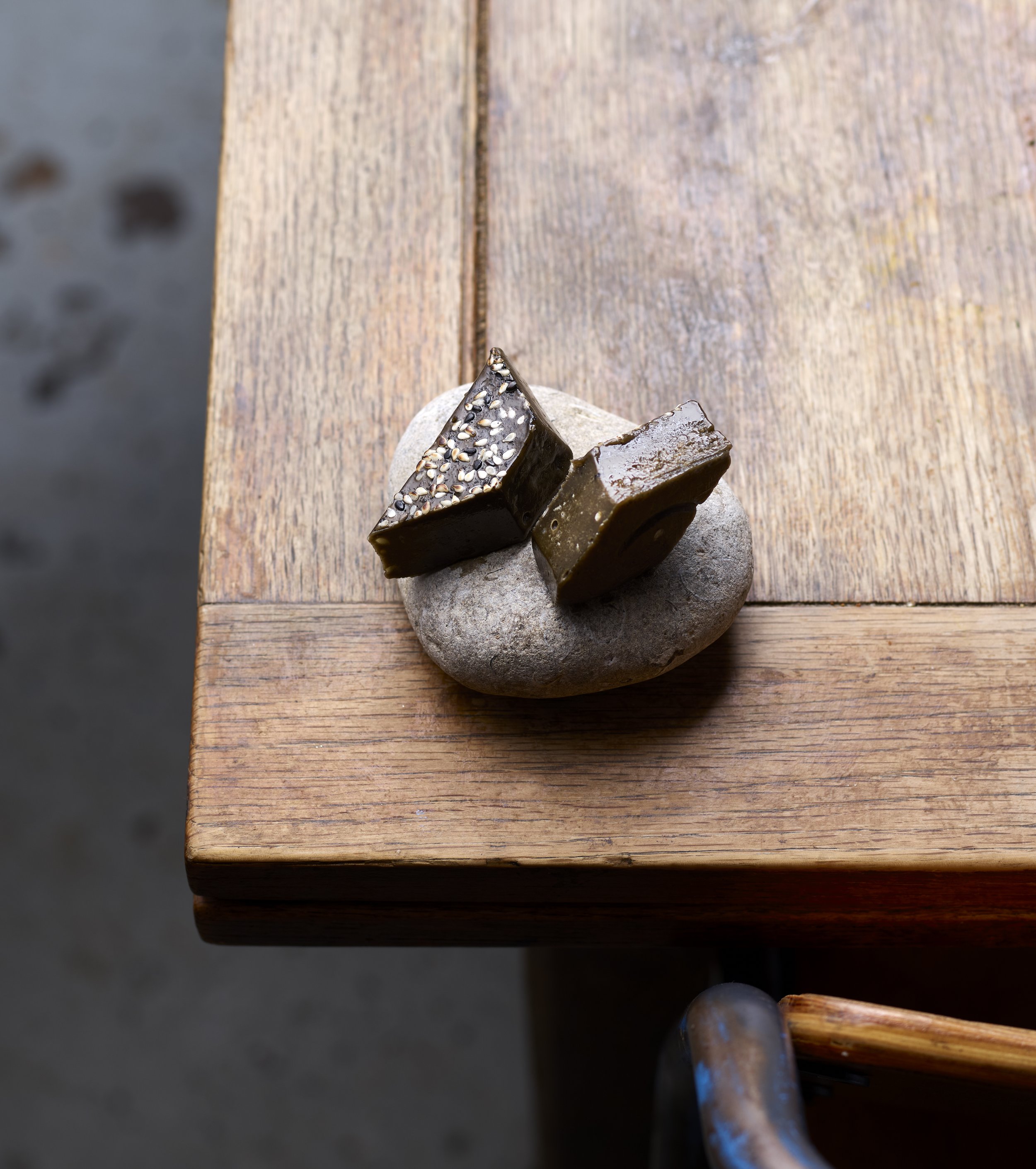 Close-up of two pieces of dark chocolate with sesame seeds on top, resting on a textured gray stone on a wooden surface.