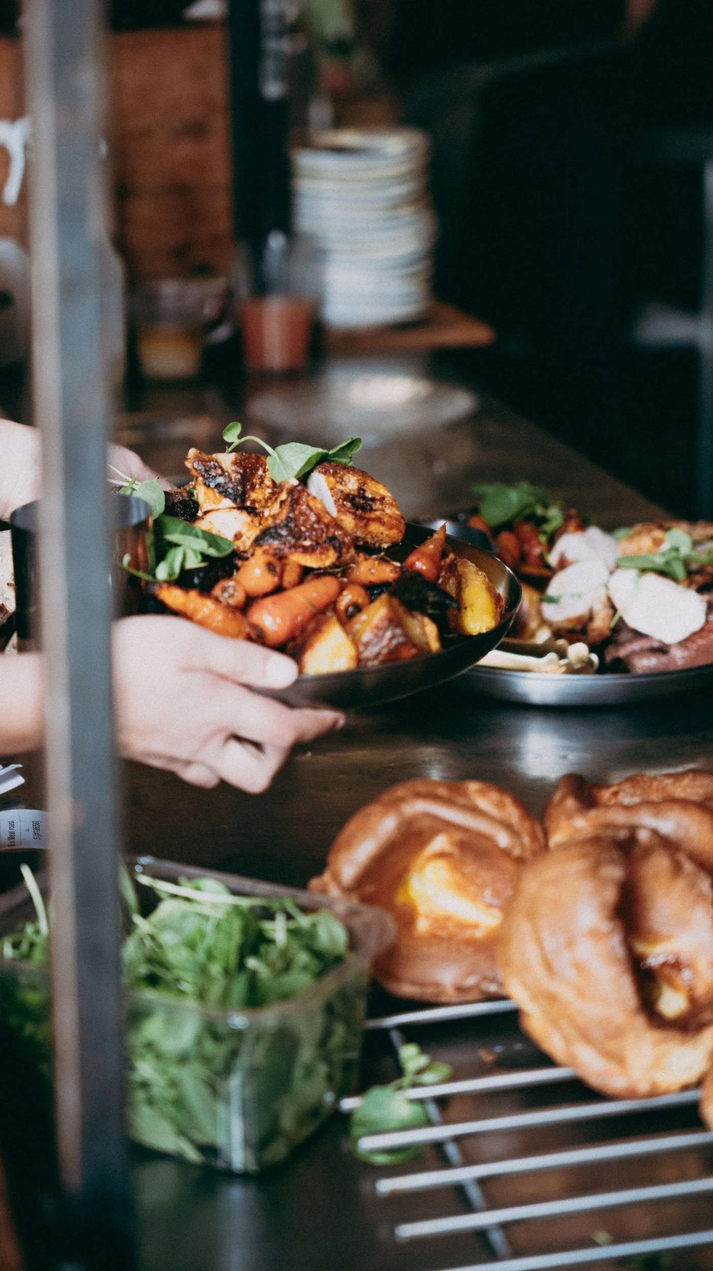 Person holding a bowl of roasted vegetables at a restaurant kitchen with plates and dishes in the background.