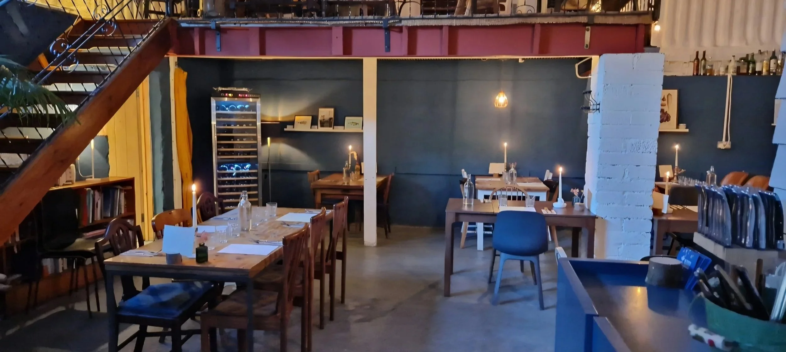 Interior of a cozy restaurant with wooden tables set for dining, candles on each table, a white brick pillar, dark blue walls, and a staircase in the upper left corner.