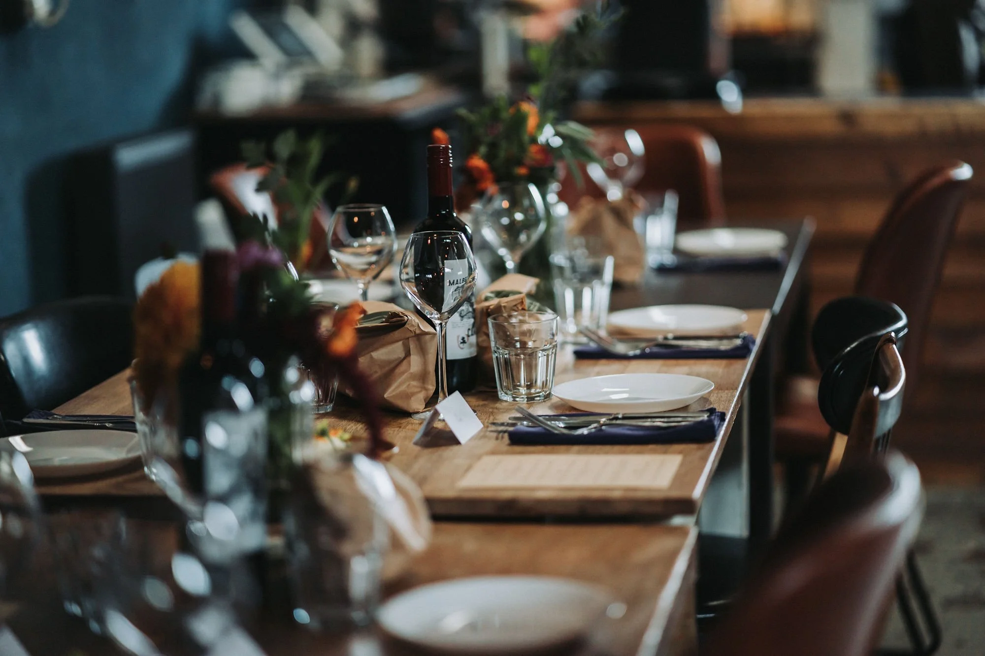 A long dining table set for a meal with wine bottles, wine glasses, and tableware, decorated with flower arrangements in a cozy, warmly lit restaurant or cafe.