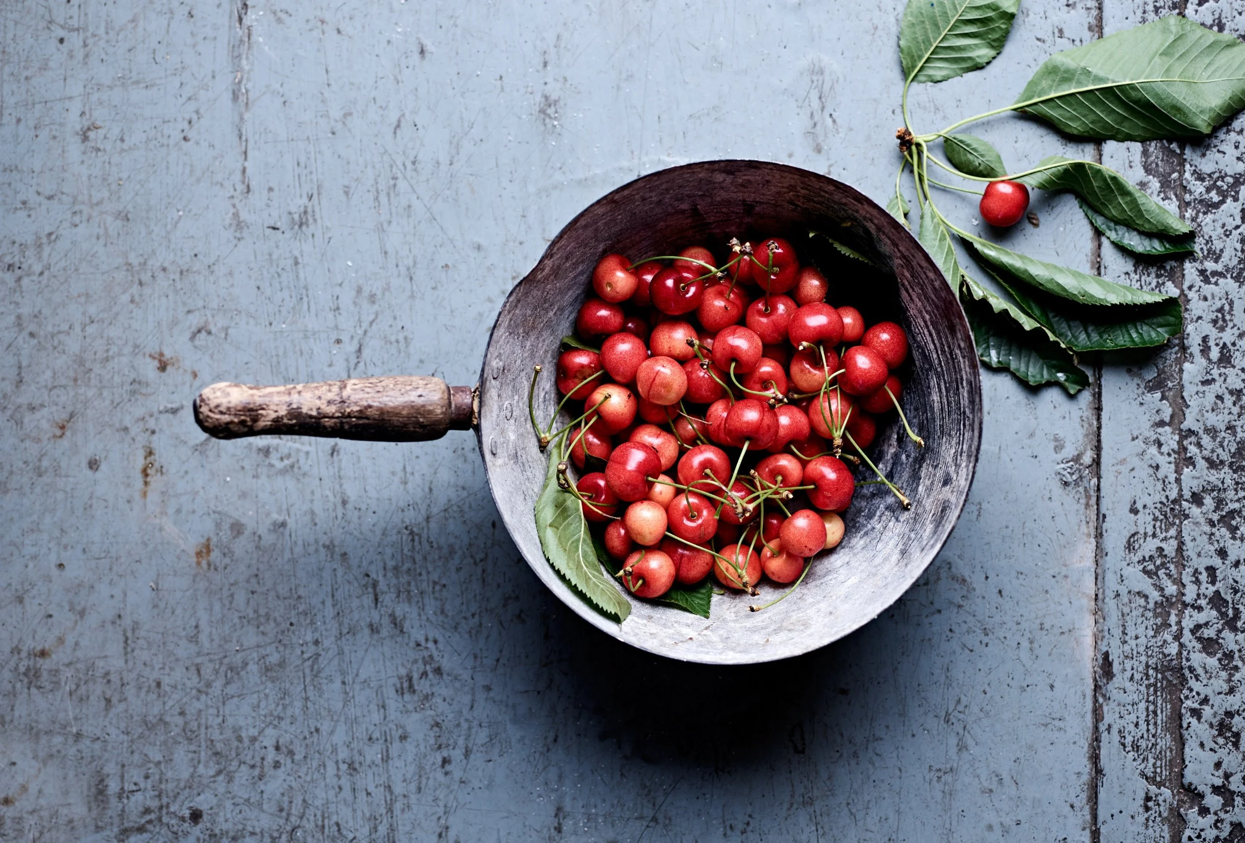 A rustic wooden bowl filled with red cherries, some with stems, placed on a weathered light blue surface with a few green cherry leaves scattered around.