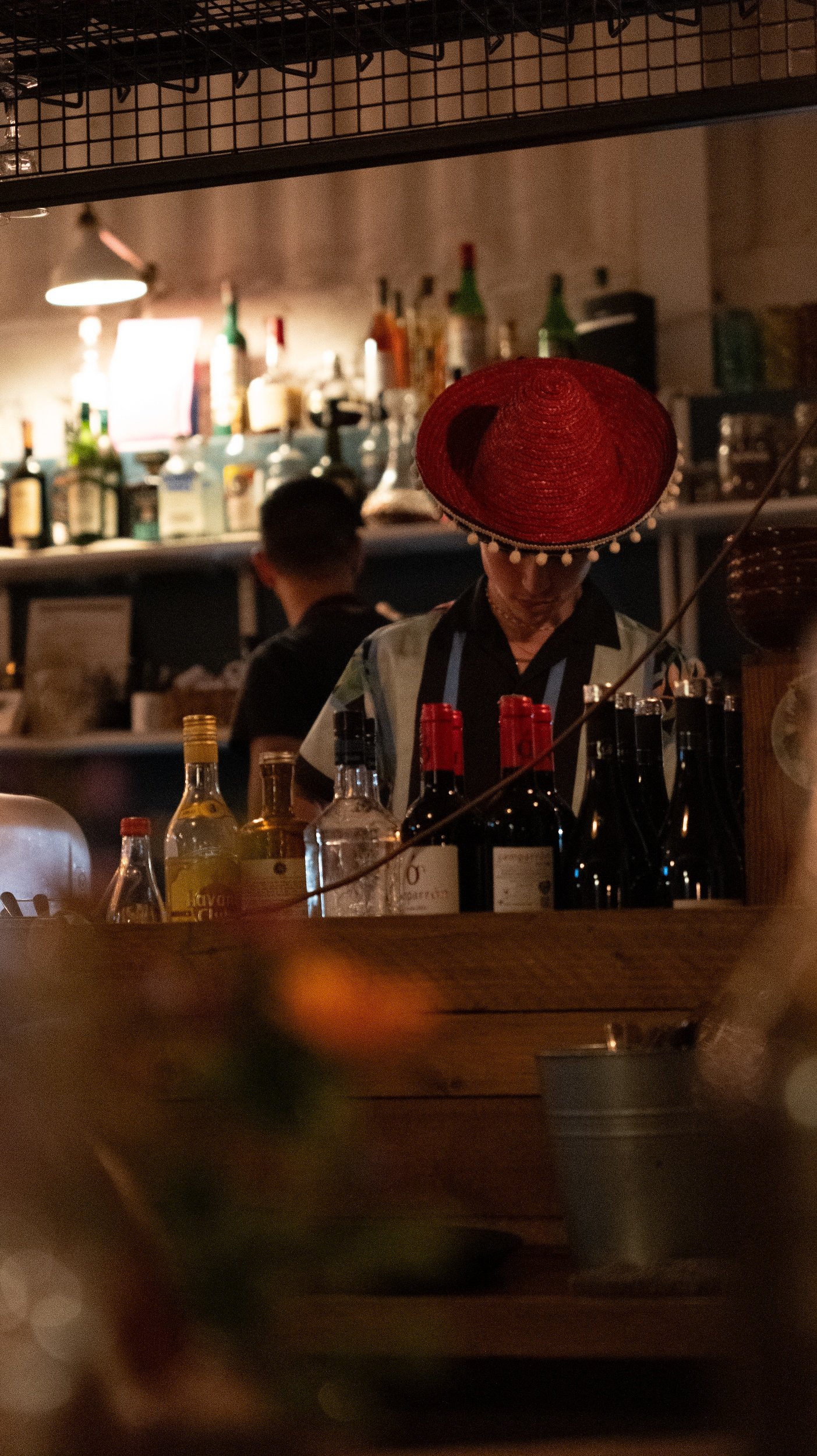 A bartender wearing a large red hat with white pom-pom trim preparing drinks behind a bar with various bottles in the background.