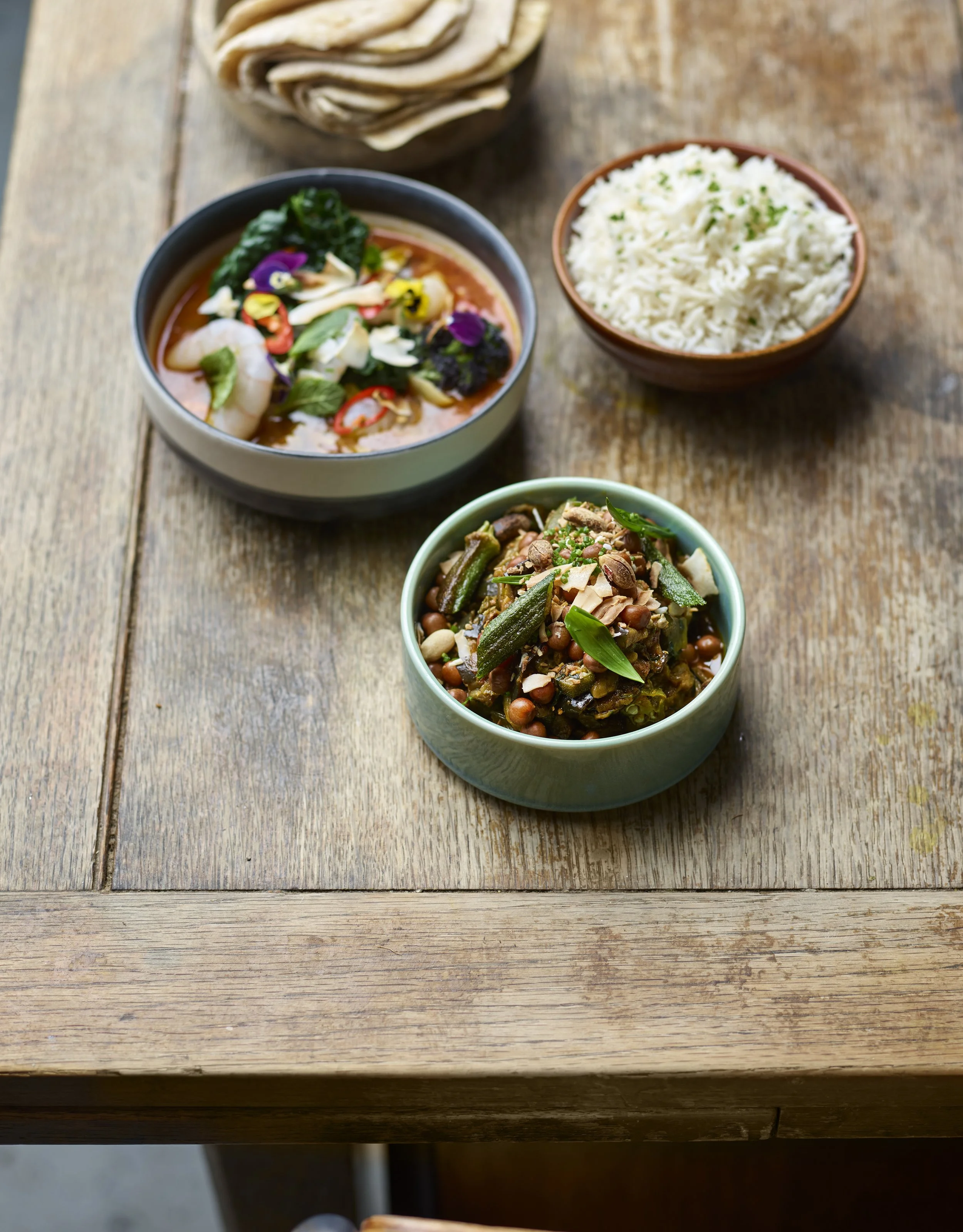 Assorted Asian dishes on a wooden table, including a bowl of spicy shrimp and vegetable soup, a bowl of cooked white rice, and a bowl of vegetable stir-fry with green beans and almonds.