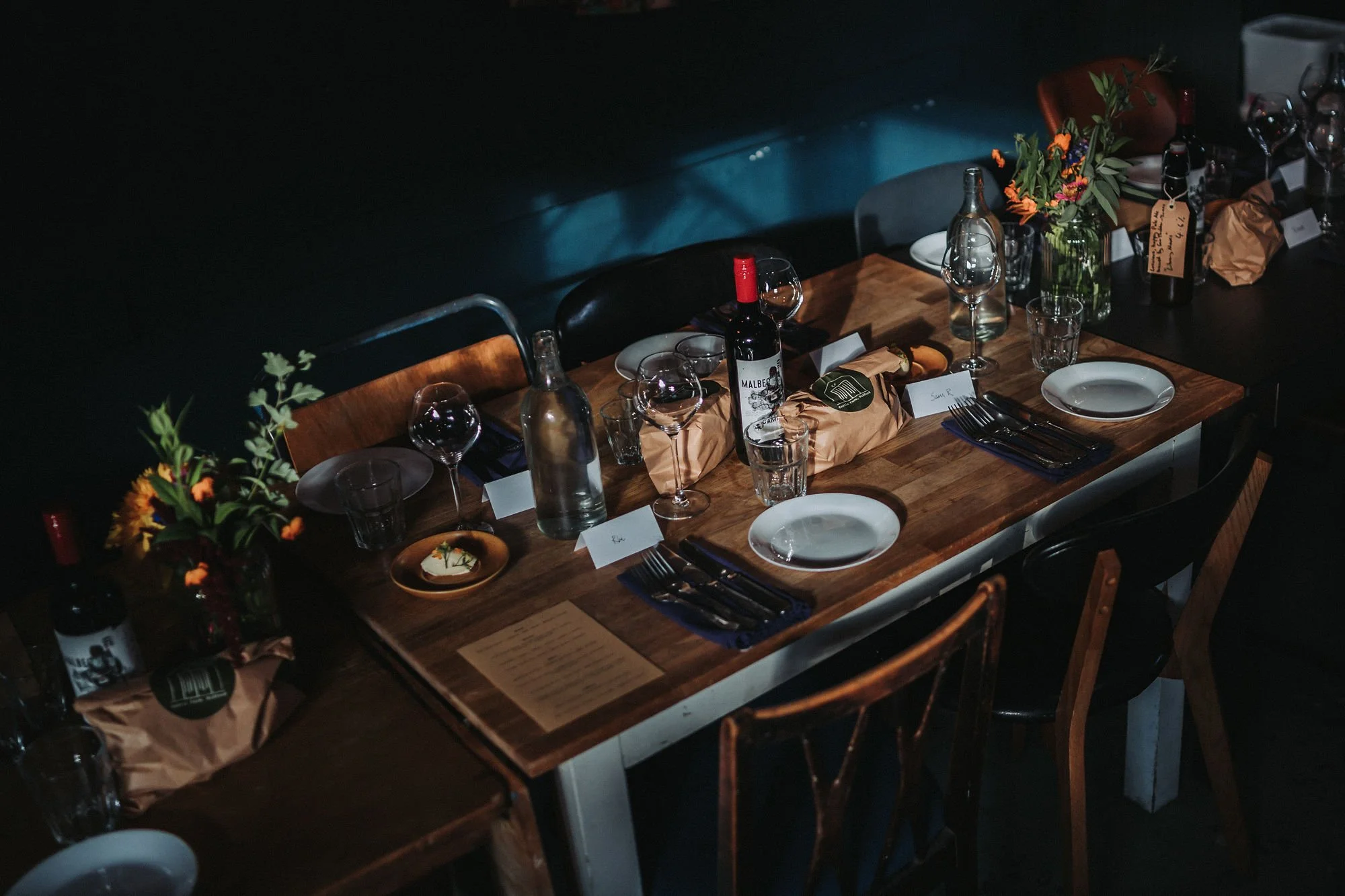 A dimly lit dining table set for a meal, decorated with flowers in vases, bottles of wine, glasses, plates, and utensils.