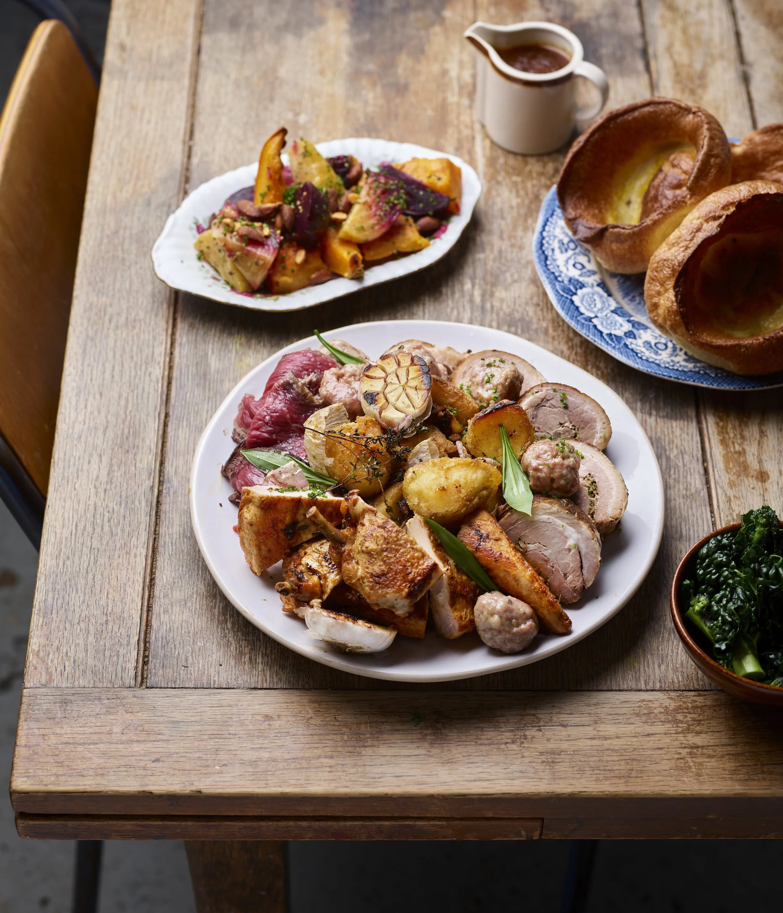 Assorted roasted meats with herbs and vegetables, garnished with fresh herbs on a white plate, accompanied by Yorkshire puddings, served on a rustic wooden table.