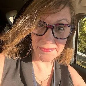 A woman with shoulder-length hair and glasses taking a selfie in a car, smiling at the camera.