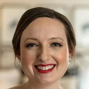 Close-up of a smiling woman with brown hair and red lipstick in a bright indoor setting.