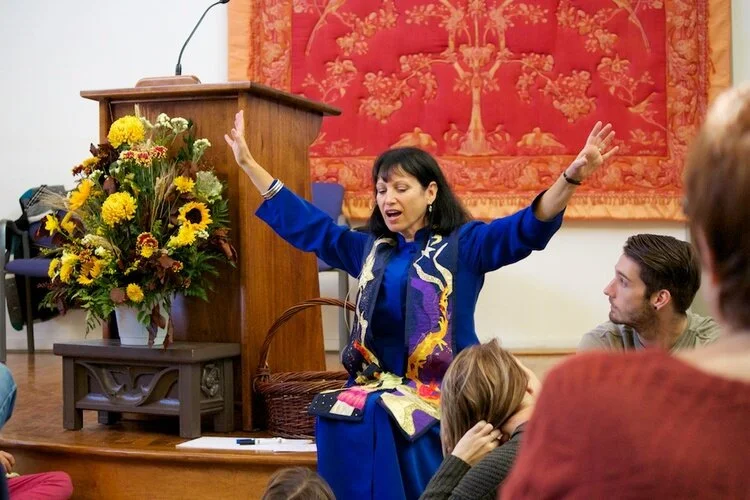 Rev. Diane dressed in blue, holding her hands up while reading.
