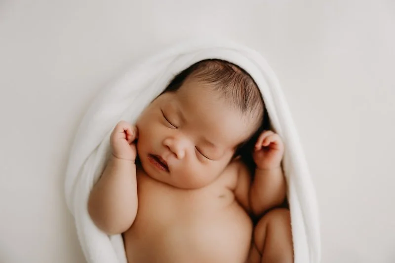A sleeping newborn baby wrapped in a white towel, lying on a white surface.