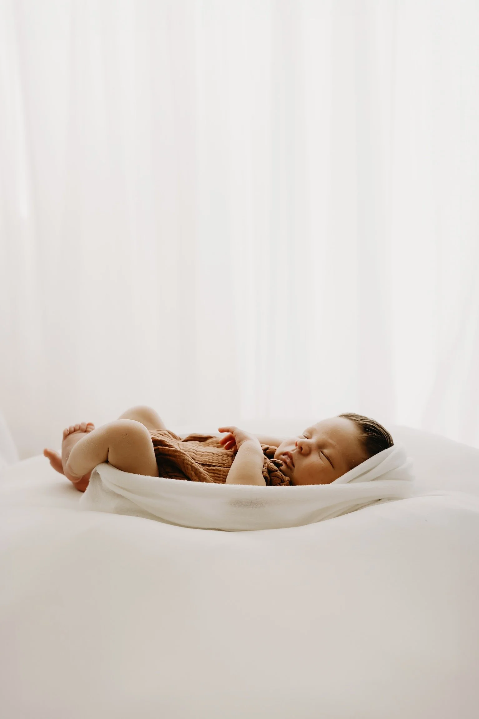 A young baby sleeping peacefully on a bed wrapped in a white blanket, wearing a brown outfit, with a white pillow under the head and a white sheet covering part of the body.