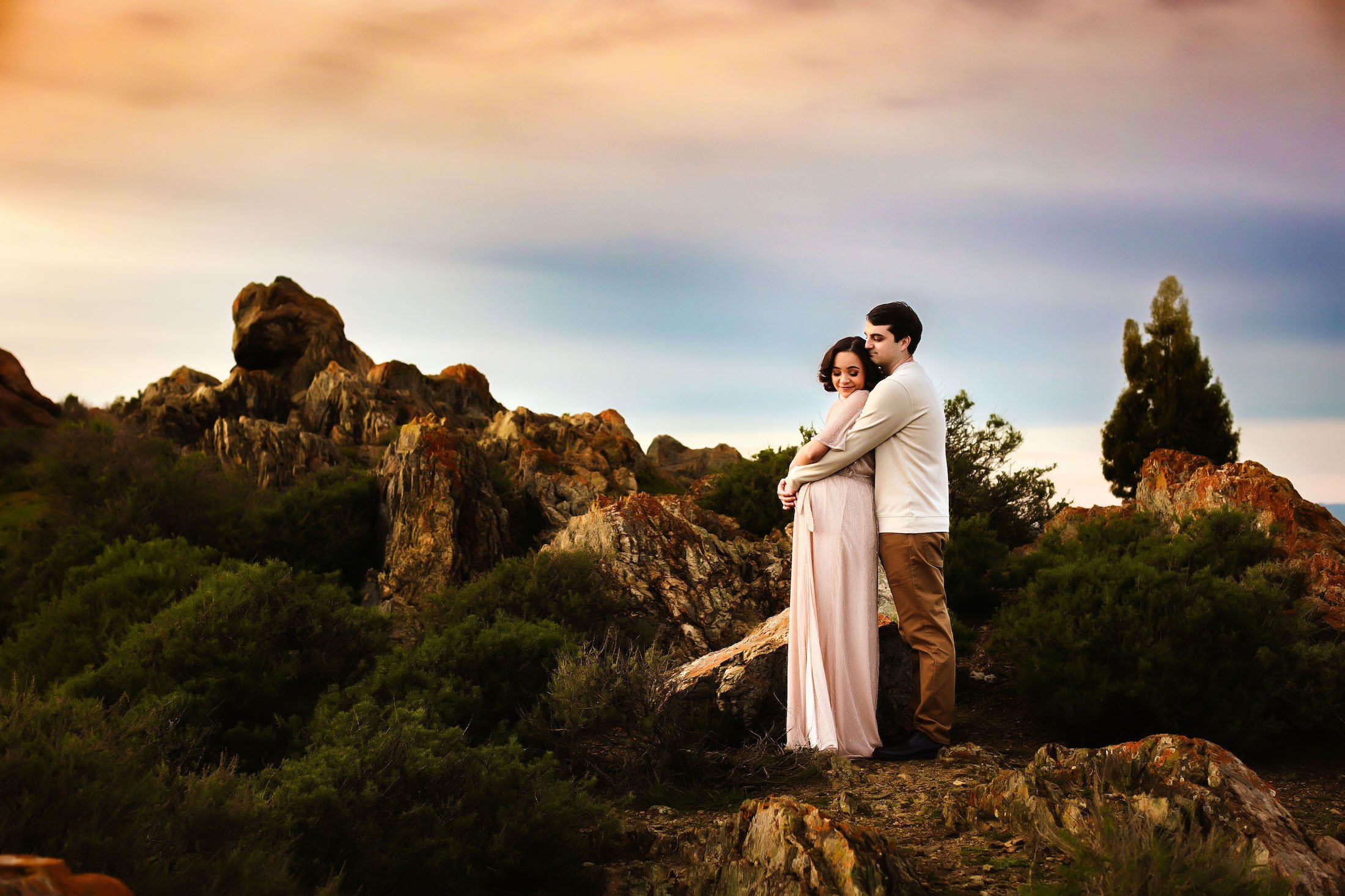 A couple standing close together on rocky terrain with a dramatic sky overhead, mountains and trees in the background, embracing each other.