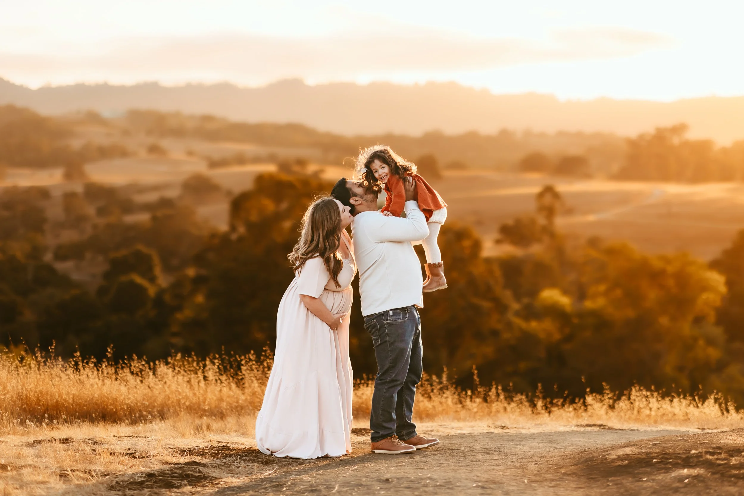 A family of three standing outdoors during sunset, with a scenic landscape of hills and trees in the background. The father is lifting the daughter into the air while the mother stands beside them, all smiling, in a Golden Hour maternity photoshoot. 