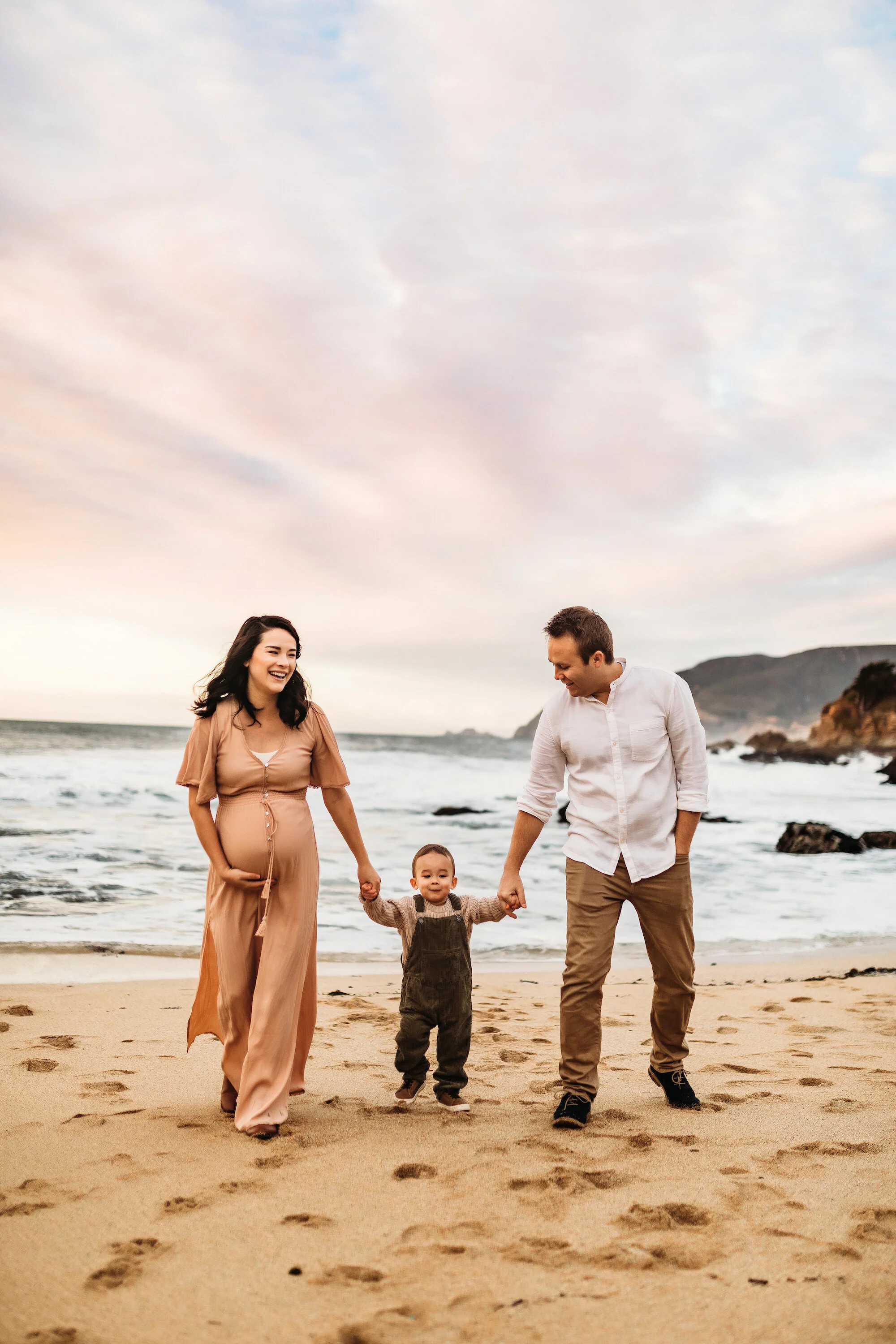 A family of four walking hand in hand along the beach with sand, ocean, and cloudy sky in the background during sunset.