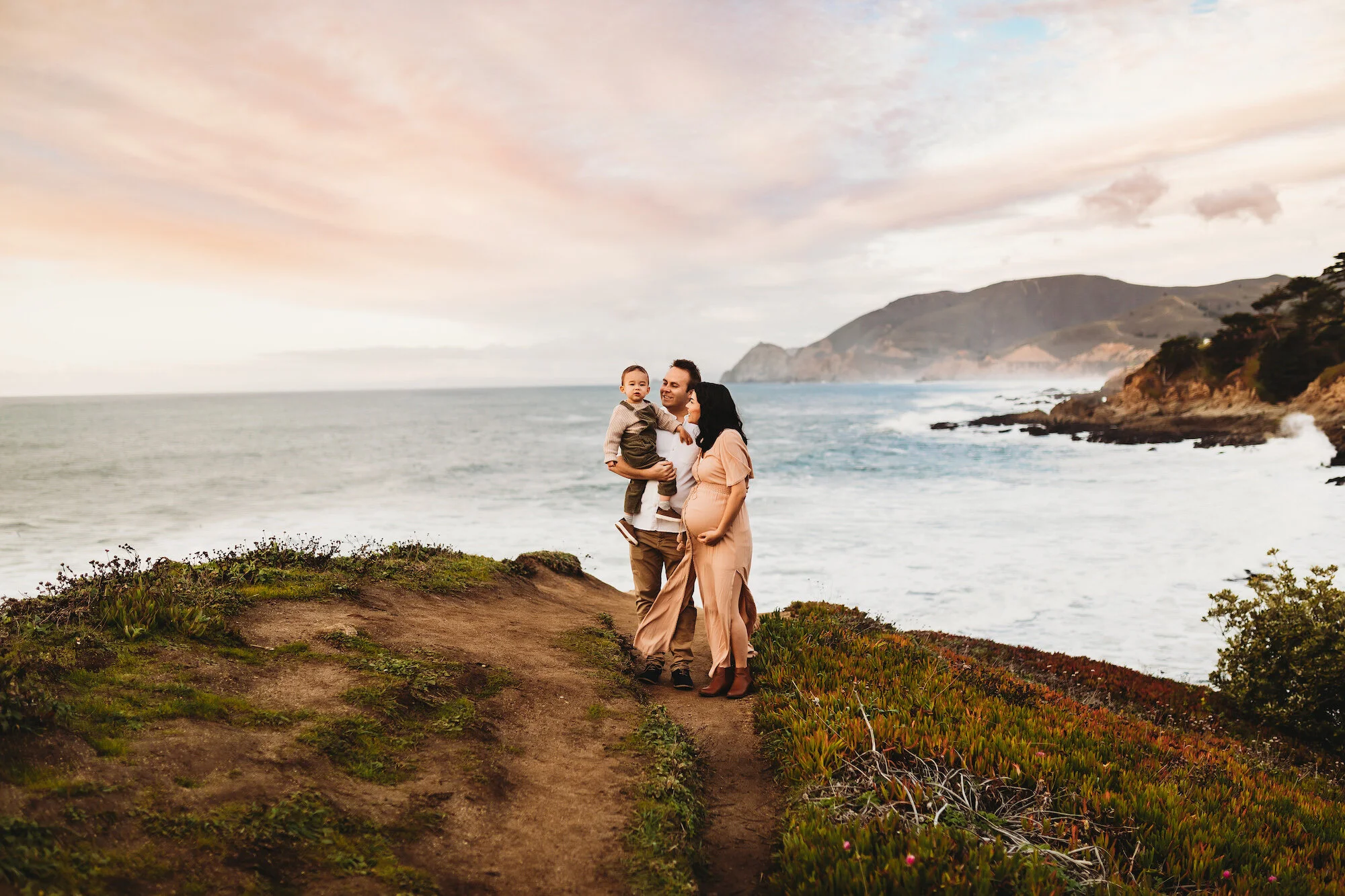 Family of three walking along a coastal path during sunset, with ocean and mountains in the background.