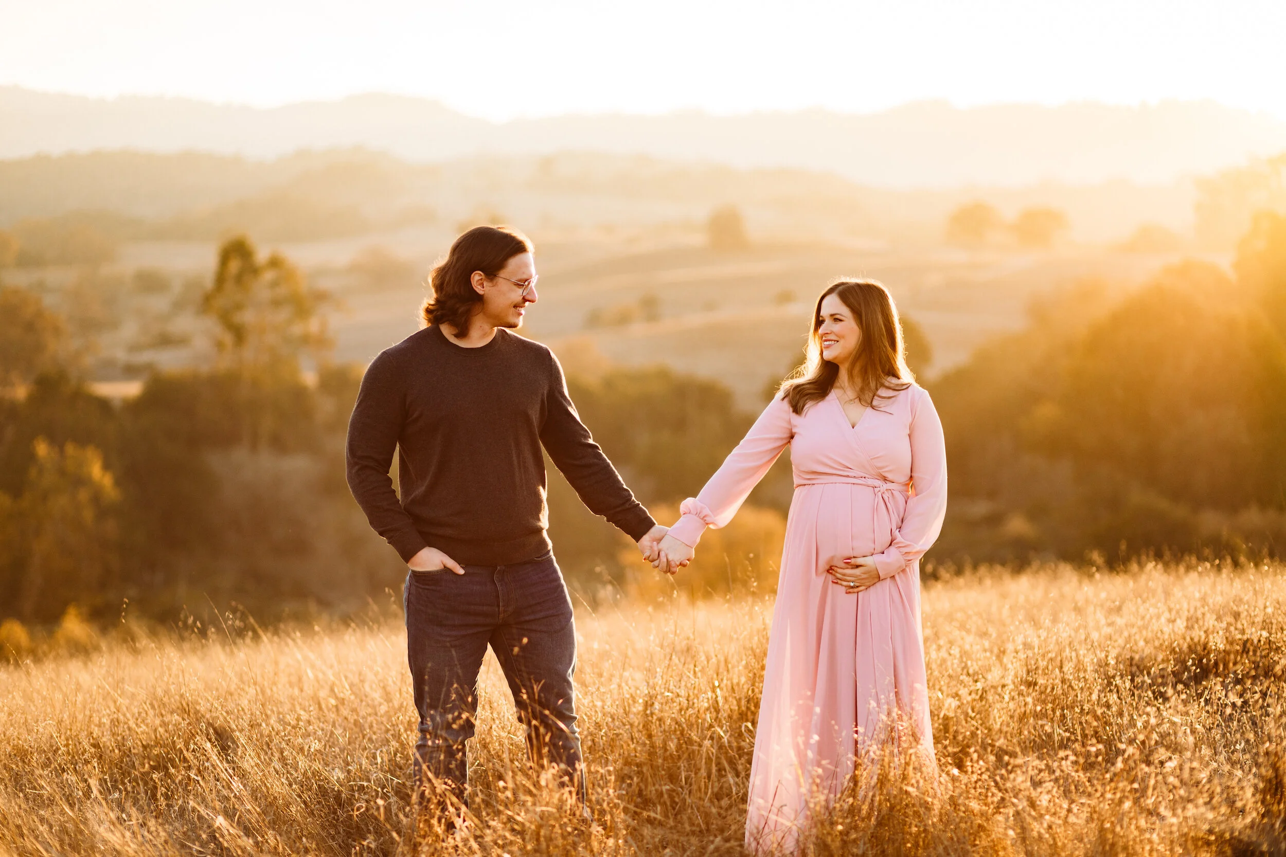 A pregnant woman in a pink dress holding her belly and a man in casual clothing holding hands and smiling at each other in a field at sunset.