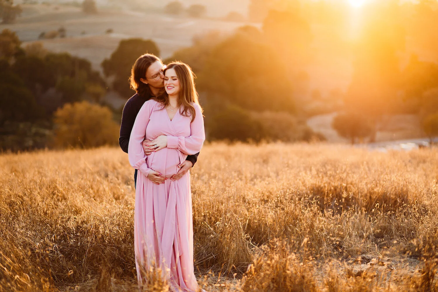 A couple standing in a field of golden grass during sunset, with the woman holding her pregnant belly and smiling, while the man embraces her from behind and kisses her on the head.