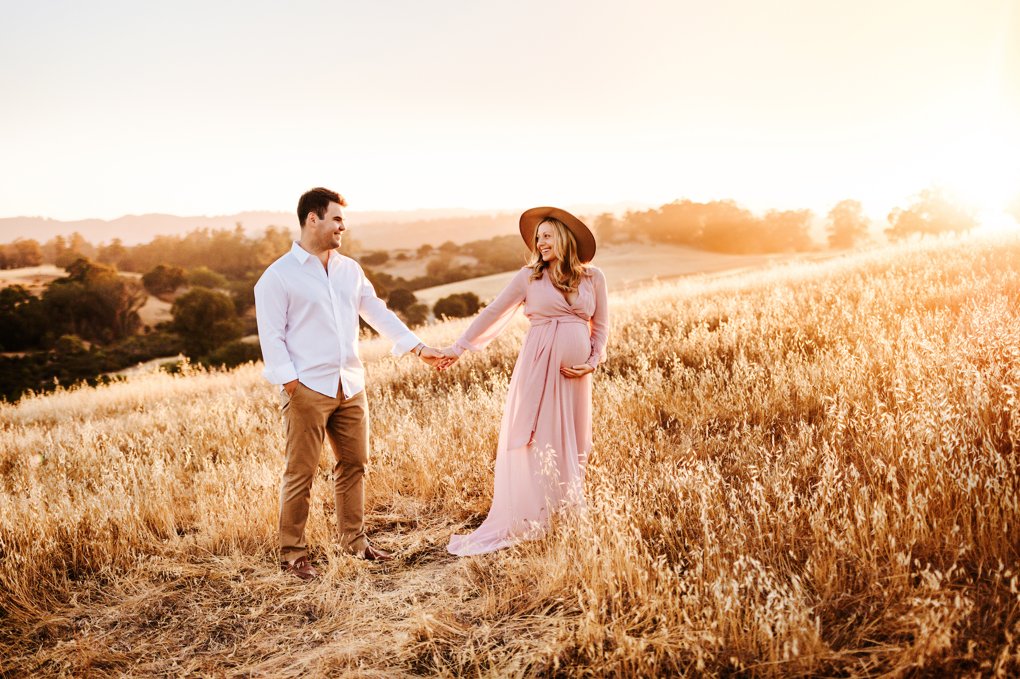 A couple holding hands in a field during sunset. The woman is pregnant, wearing a pink dress and a wide-brimmed hat. The man is in a white shirt and khakis. Hills and trees are visible in the background.
