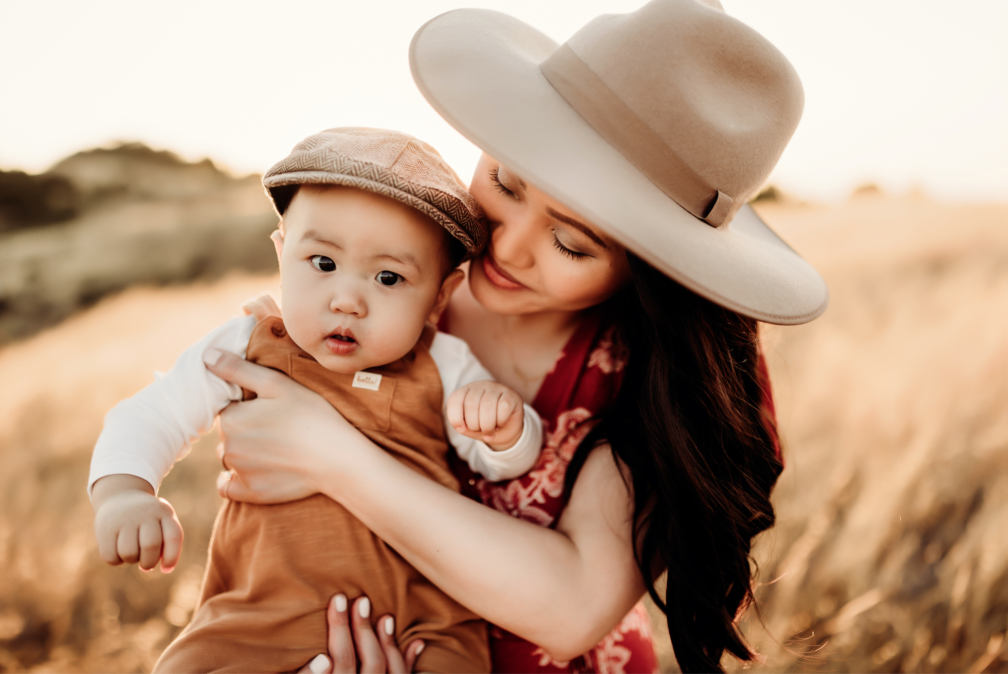 A woman with long dark hair in a large beige hat holding a young child with a brown cap outdoors at sunset.